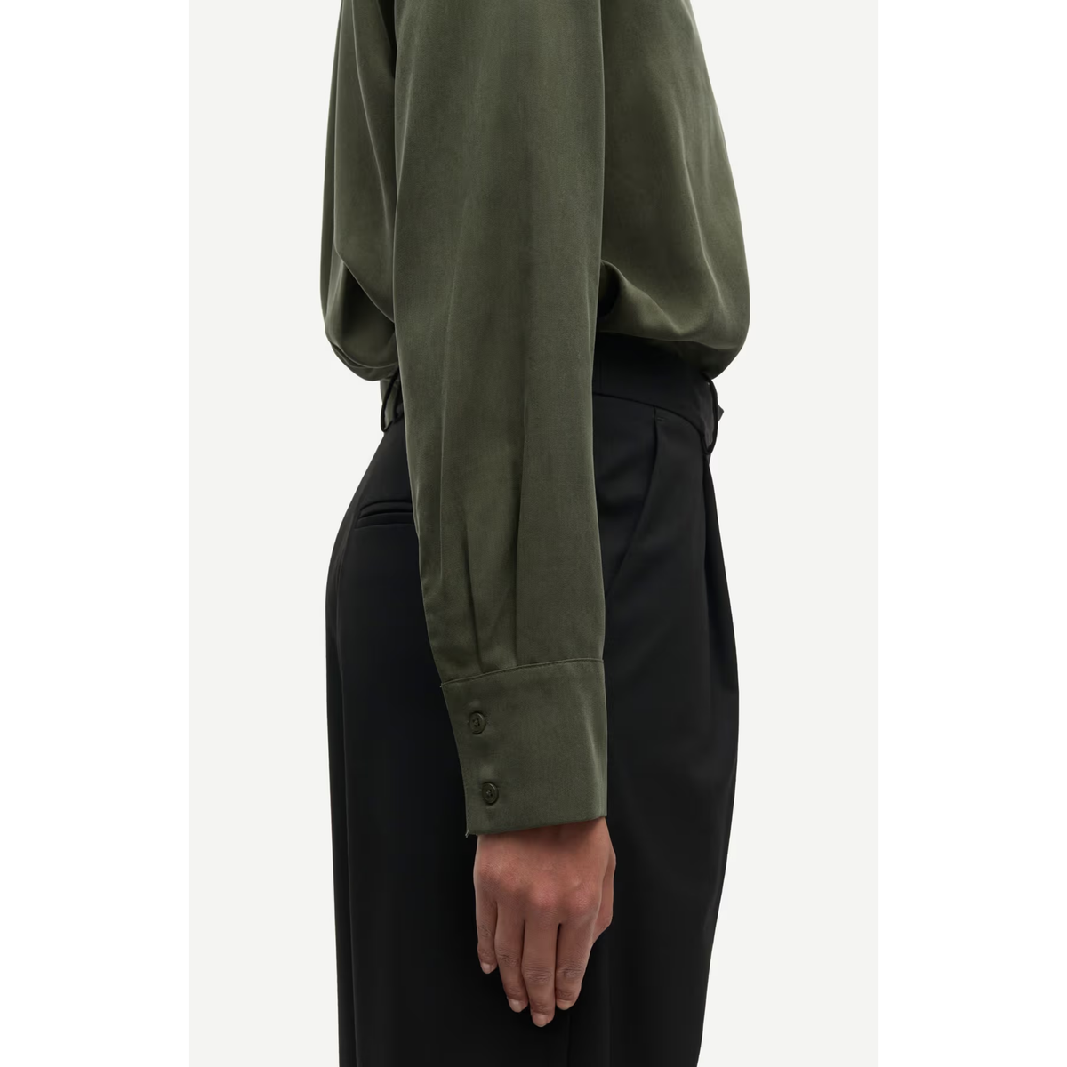 A side-profile view of a person wearing an olive green long-sleeve blouse with buttoned cuffs and black trousers, shown against a plain light background.