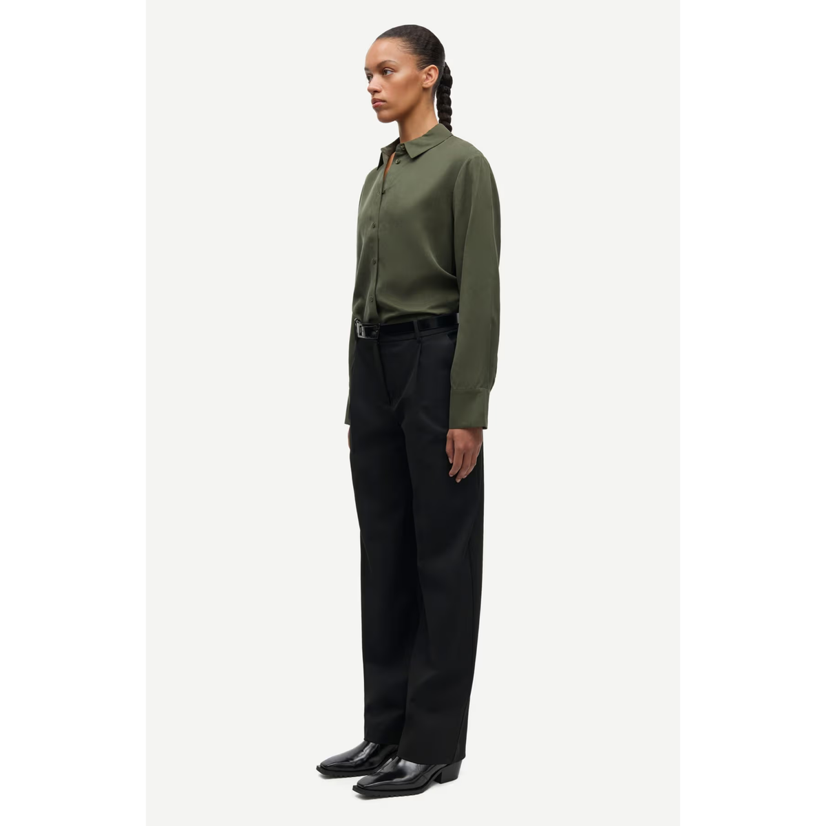 A woman stands in profile wearing a long-sleeve olive green button-up shirt tucked into black trousers with black leather shoes, against a plain light gray studio background.