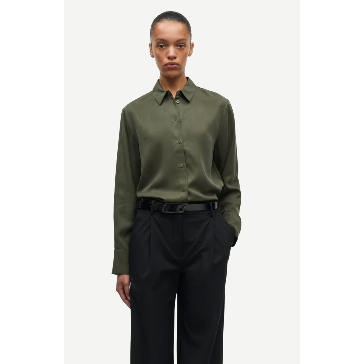 A model wearing an olive green long-sleeve button-up shirt tucked into black trousers with a black belt, standing against a light gray studio background.