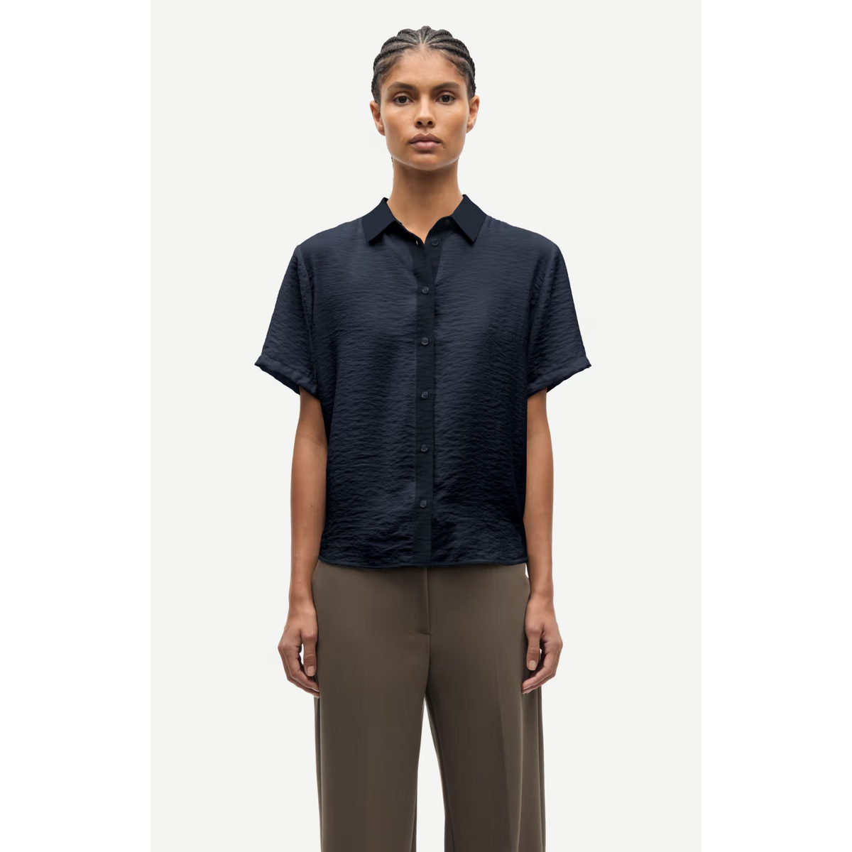A model wearing a short-sleeve button-up shirt in dark navy blue with a subtle crinkled texture, paired with olive brown trousers, set against a plain light gray background.