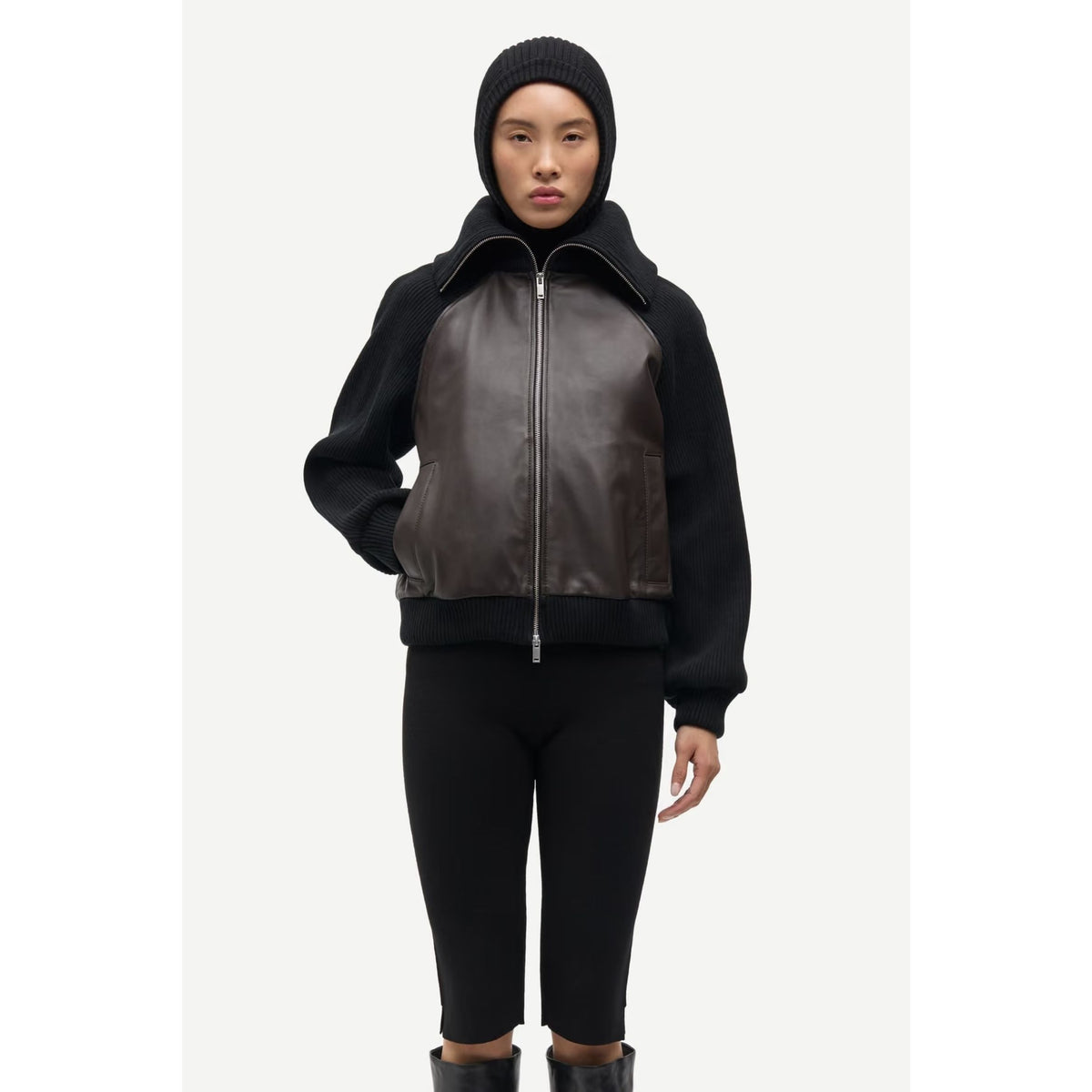 A model wearing a black and dark brown leather jacket with a high collar, black knit beanie, black leggings, and black boots, standing against a light gray studio background.