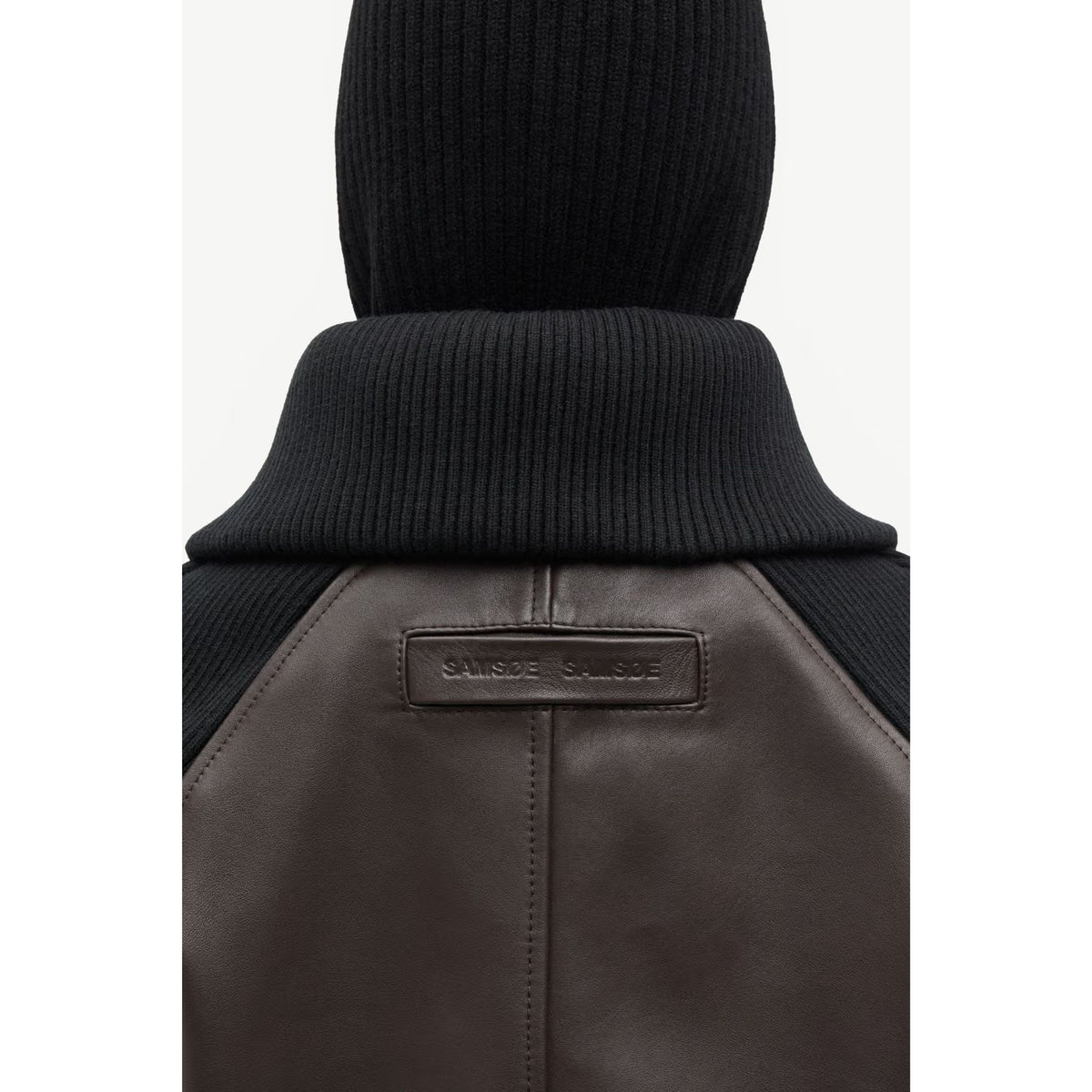 Close-up of the back of a dark brown leather jacket with a black ribbed knit collar and hood, featuring a small 'SAUNDERSSALUTE' brand patch on the leather.