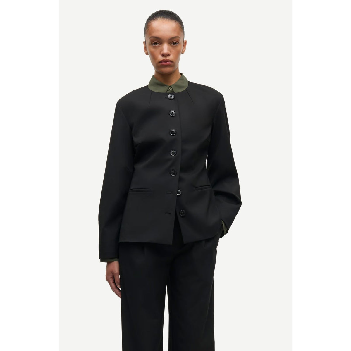 A model wearing a black structured jacket with a contrasting olive green collar and matching black trousers, photographed against a light gray studio background.
