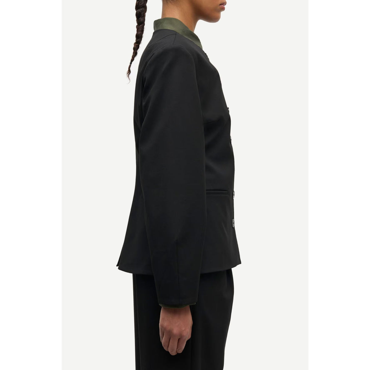 Side profile of a model wearing a black long-sleeve blazer with an olive green collar, paired with black trousers, against a plain off-white background.