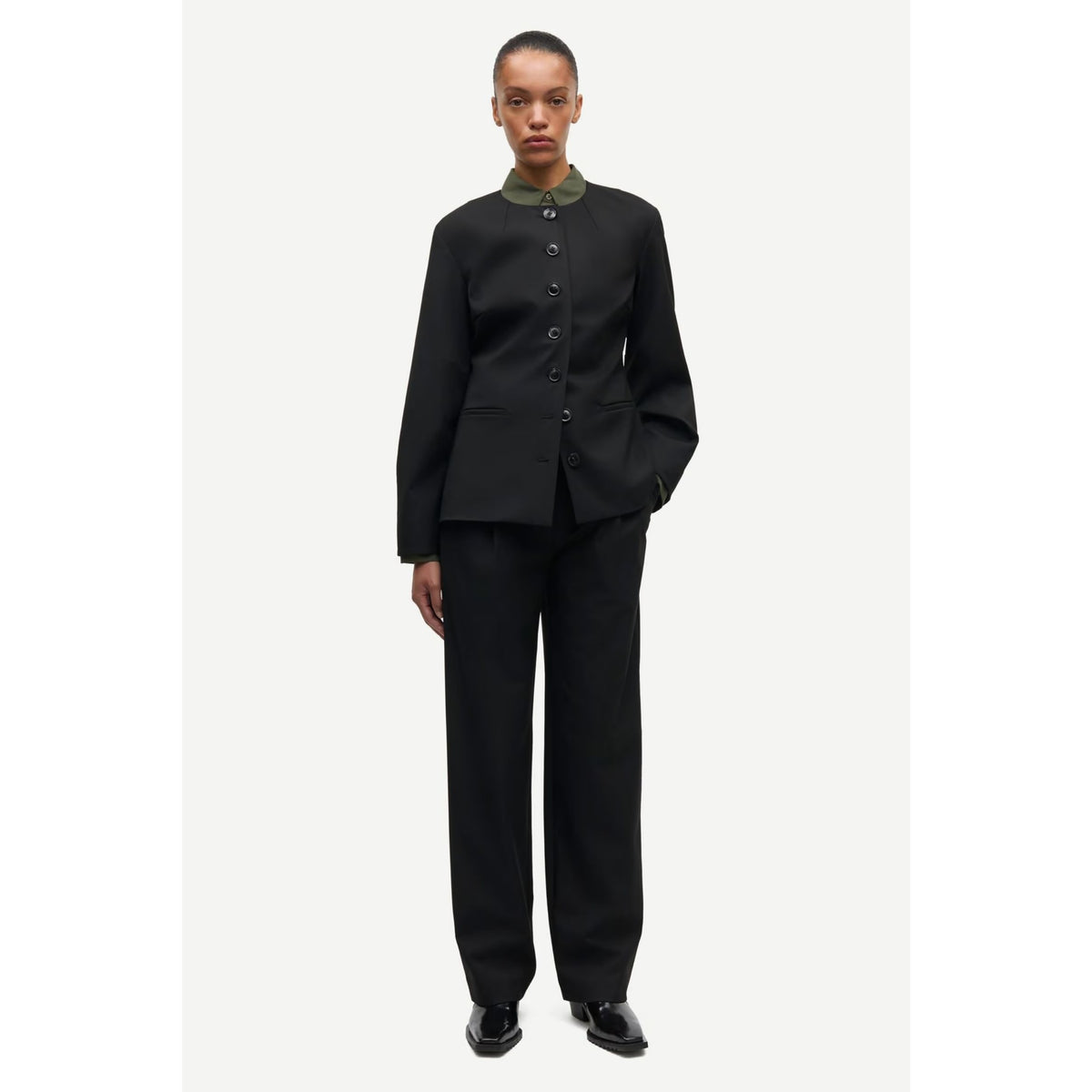 A model wearing a black tailored suit with a structured jacket featuring a high collar and front buttons, paired with matching wide-leg trousers, on a light gray background.