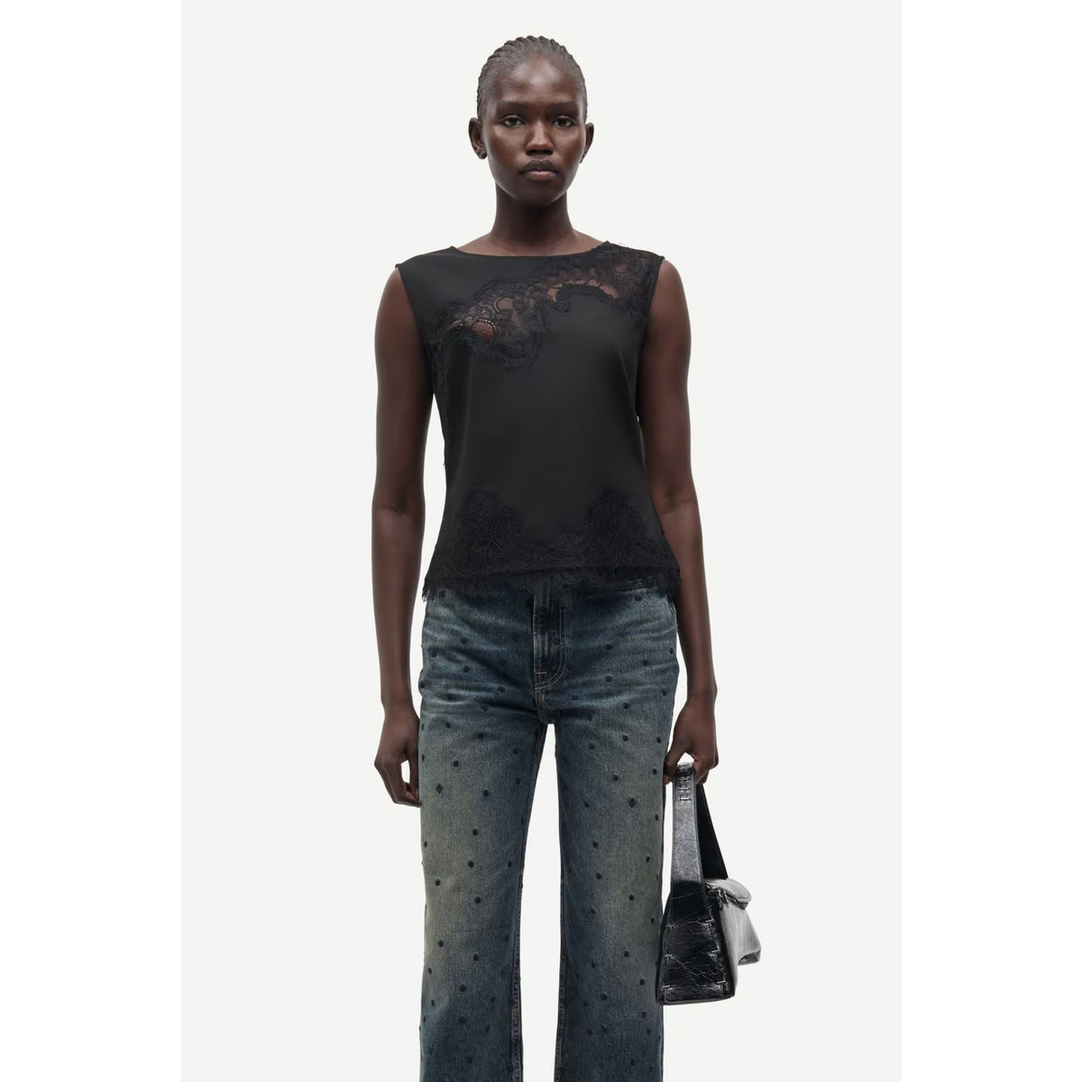 A model wearing a black sleeveless top with sheer lace detailing across the chest, paired with blue jeans featuring small black dot patterns, holding a black handbag against a light gray background.