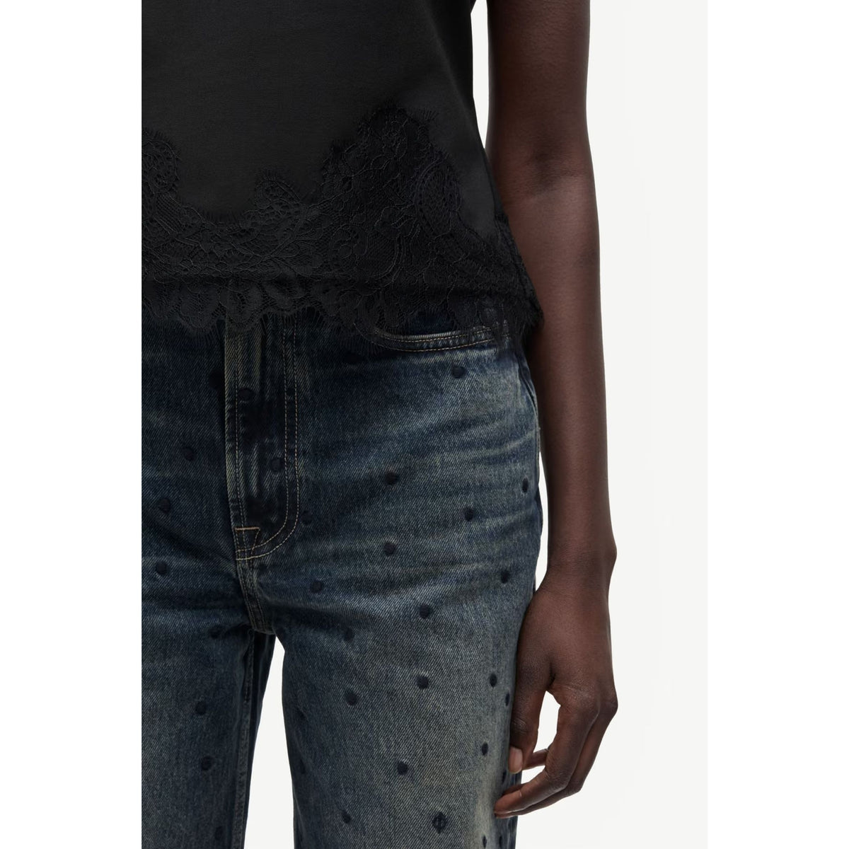 Close-up of a model wearing dark blue denim jeans with small embroidered dots and a black top with lace trim, shown against a plain light background.