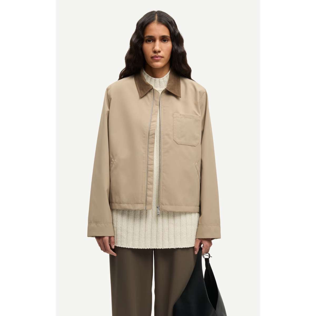 A model wearing a beige utility jacket with a zippered front and chest pocket, layered over a cream ribbed sweater and brown trousers, holding a black bag against a light gray background.