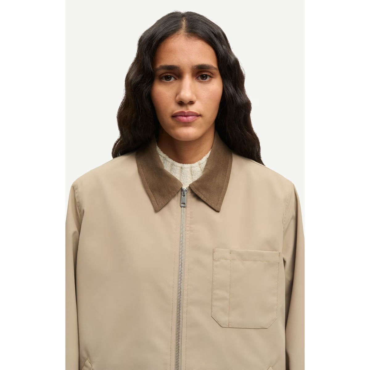 A woman wearing a beige utility jacket with a brown corduroy collar, shown from the chest up against a light gray studio background.