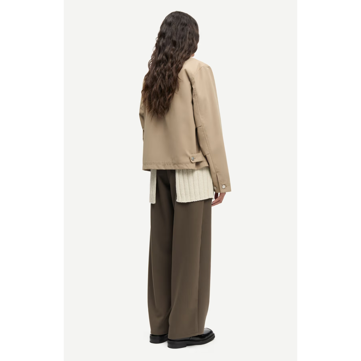 A model shown from the back wearing a beige cropped jacket over a cream ribbed top, paired with wide-leg brown trousers and black shoes, against a plain light gray background.