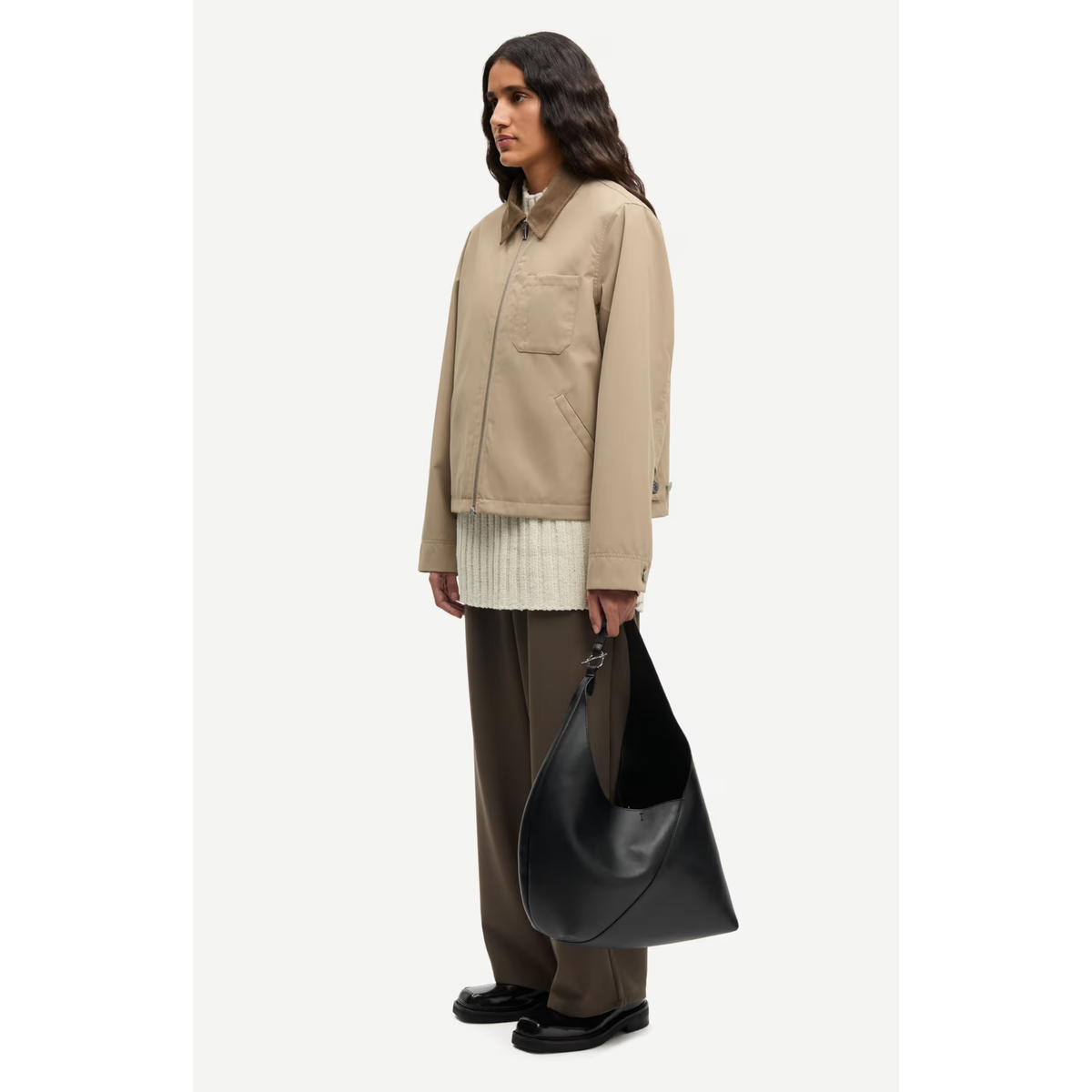 A model stands in profile wearing a beige utility jacket over a cream ribbed top, brown wide-leg trousers, and black shoes, while holding a large black leather hobo bag against a plain light background.