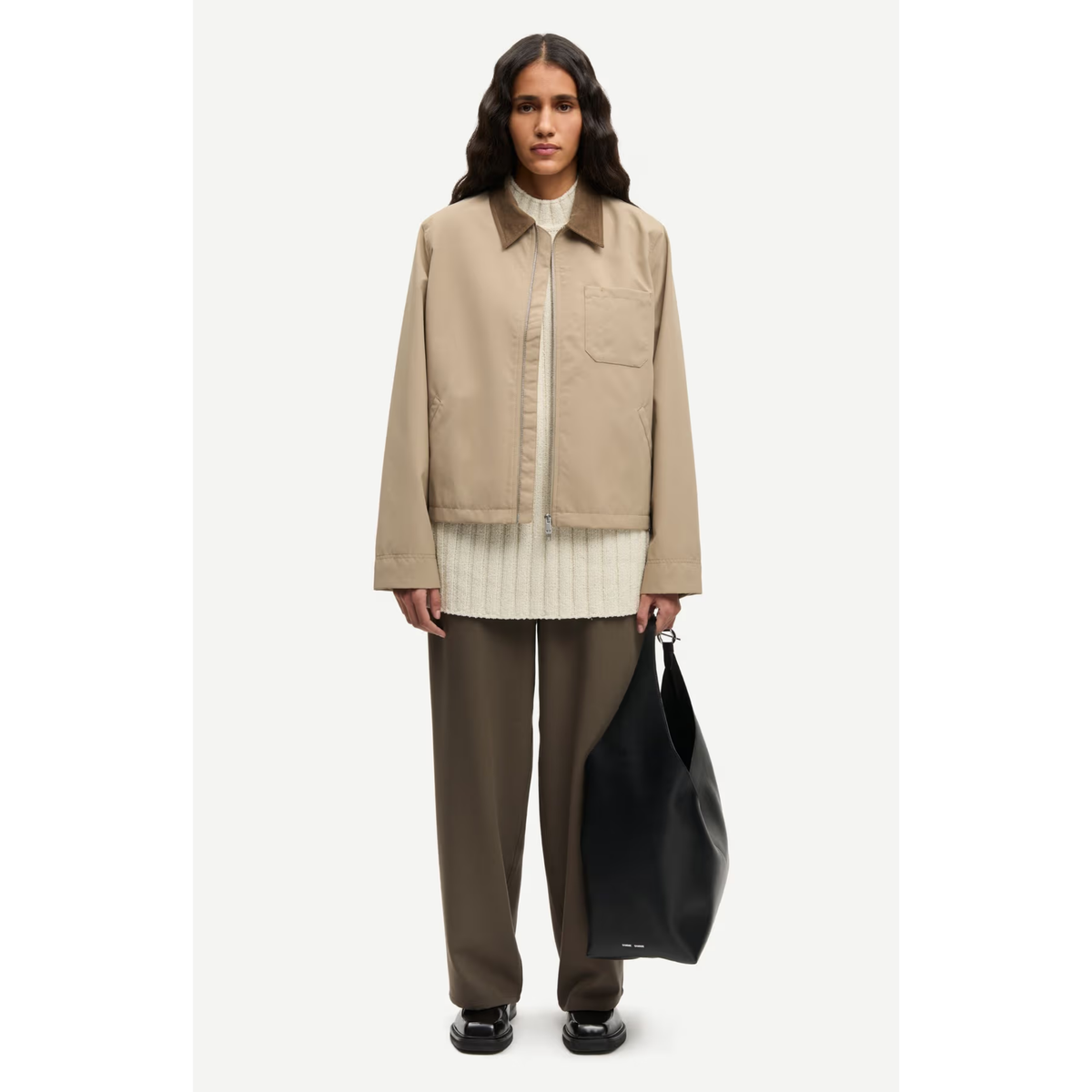 A model stands against a light gray studio background wearing a beige utility jacket over a cream ribbed shirt, olive-brown trousers, and black shoes, while holding a large black tote bag.