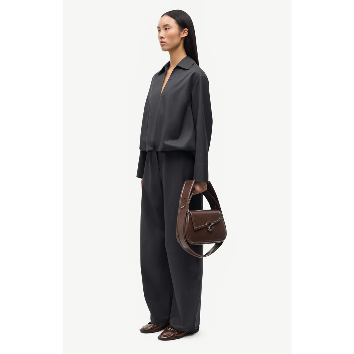 A model stands in profile wearing a dark charcoal gray long-sleeve collared shirt and matching wide-leg trousers, holding a brown leather handbag against a clean off-white studio background.