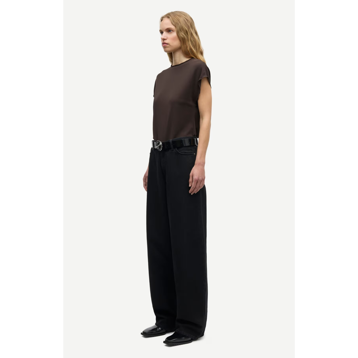 A model is shown in profile wearing a dark brown short-sleeve top, black wide-leg trousers, and a black belt with a silver buckle, set against a plain light gray background.