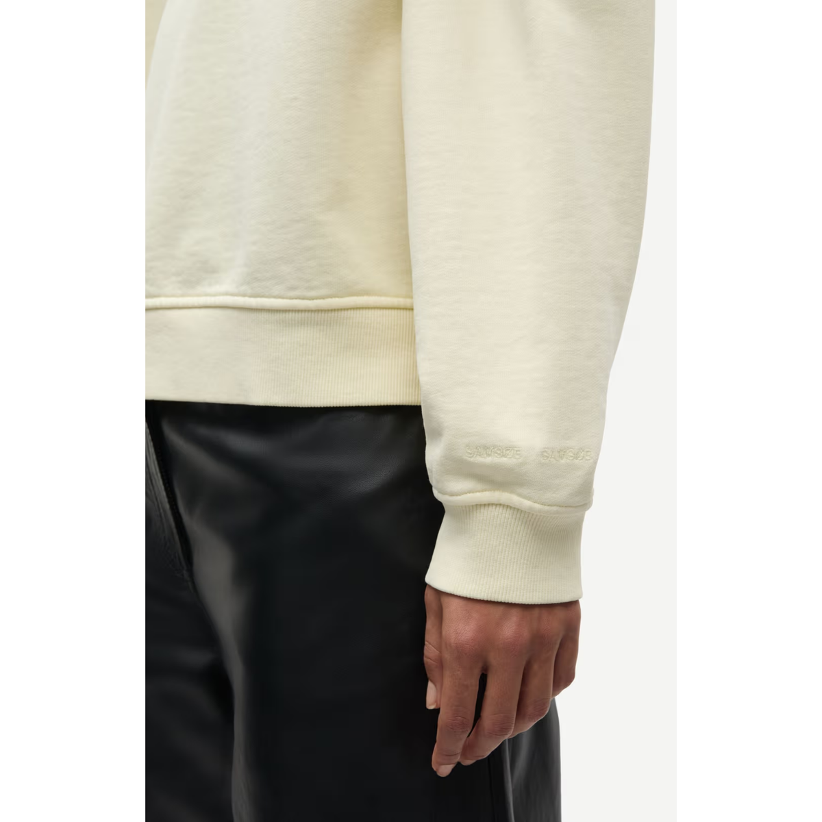 Close-up of a person wearing a cream-colored crewneck sweatshirt with ribbed cuffs and hem, paired with black pants, against a plain light background.