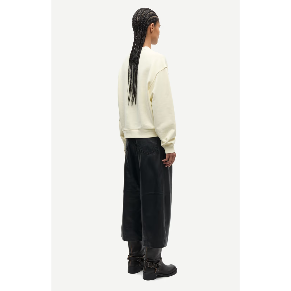 A model shown from the back wearing a cream-colored sweatshirt, wide-leg black pants, and black boots, standing against a light gray studio background.