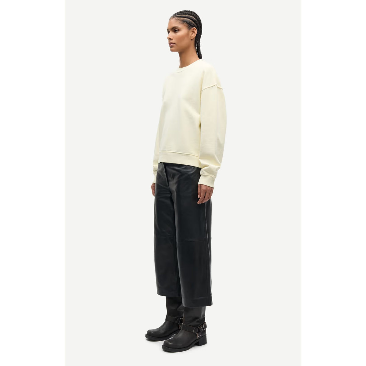 A model is shown in profile wearing a cream-colored crewneck sweatshirt, black wide-leg leather pants, and black ankle boots, set against a plain light gray studio background.