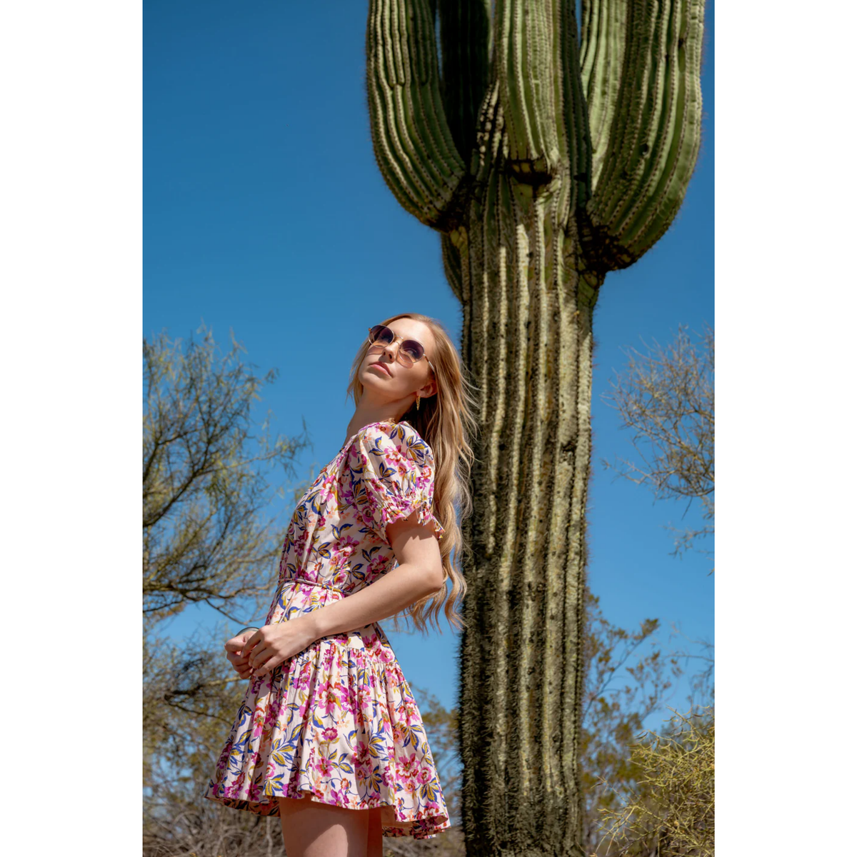 Woman in a floral dress standing next to a cactus with a clear blue sky.