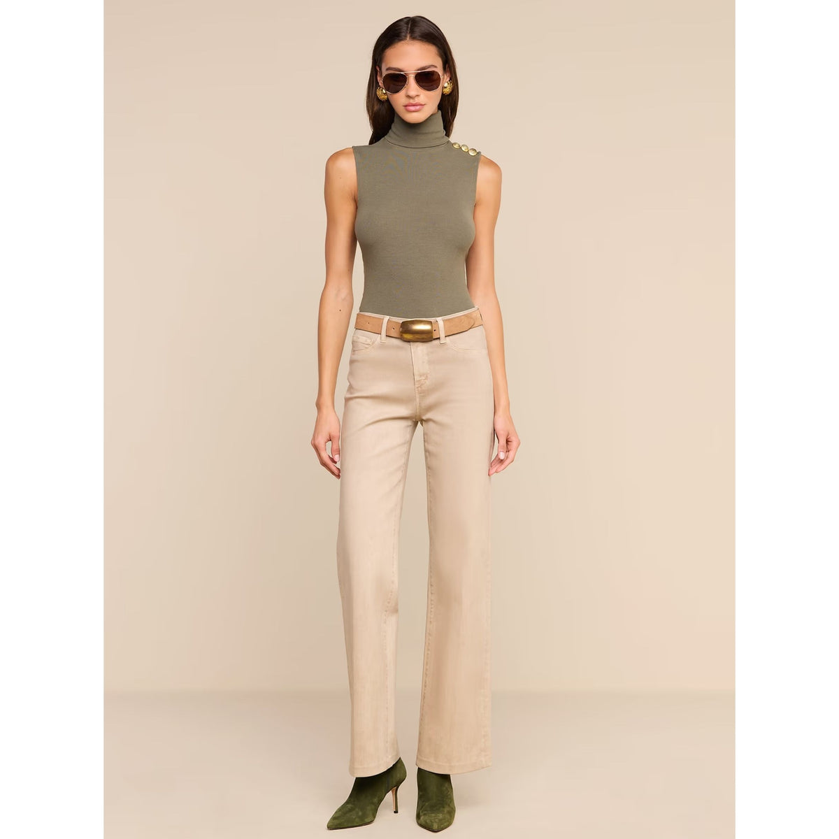 A woman modeling a sleeveless olive green mock-neck top with gold button details on the shoulder, paired with beige wide-leg pants, a tan belt, and olive green high heels against a solid beige studio background.