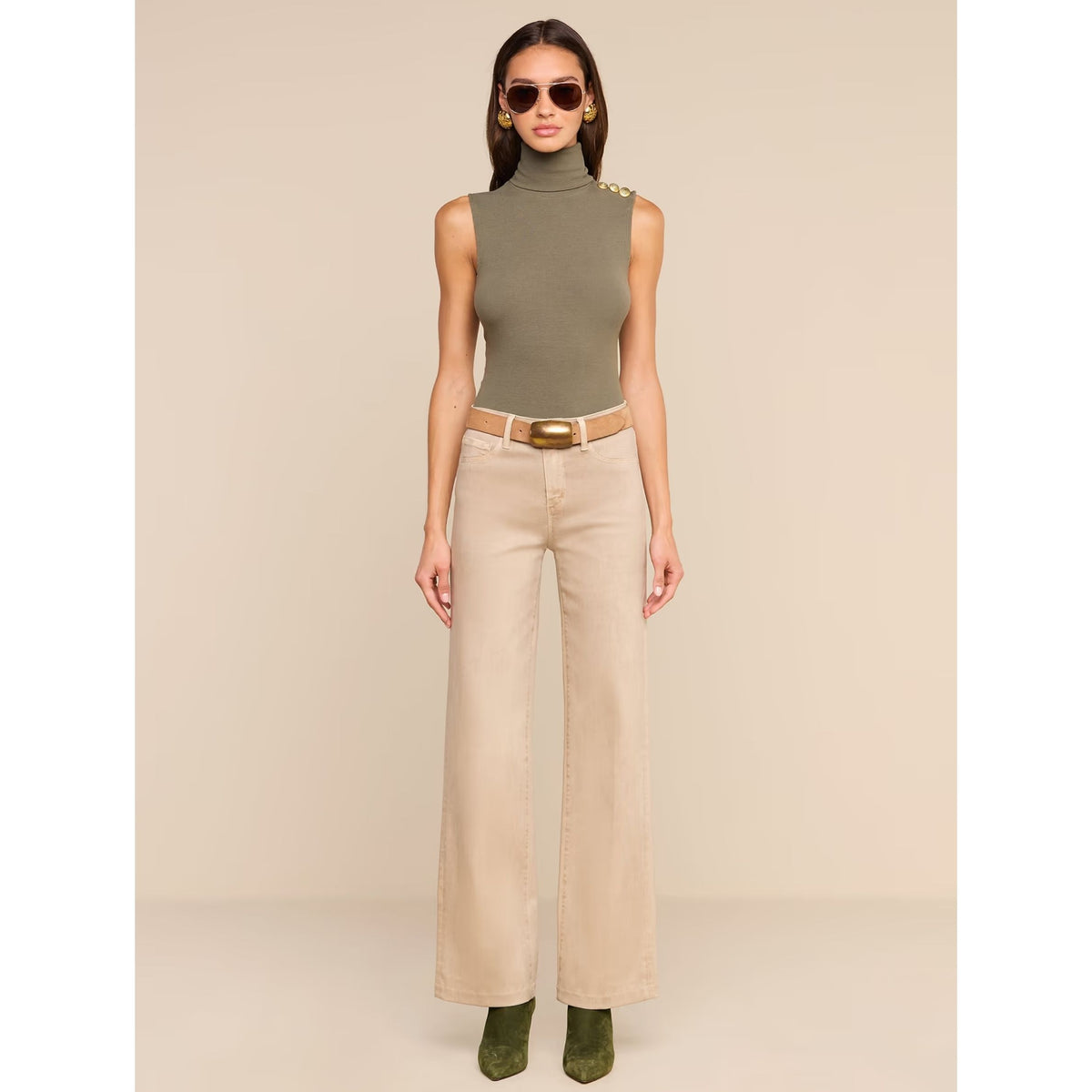 A model stands against a plain beige studio background wearing a sleeveless olive green mock-neck top with gold shoulder buttons, beige wide-leg pants with a tan belt, and olive green boots.