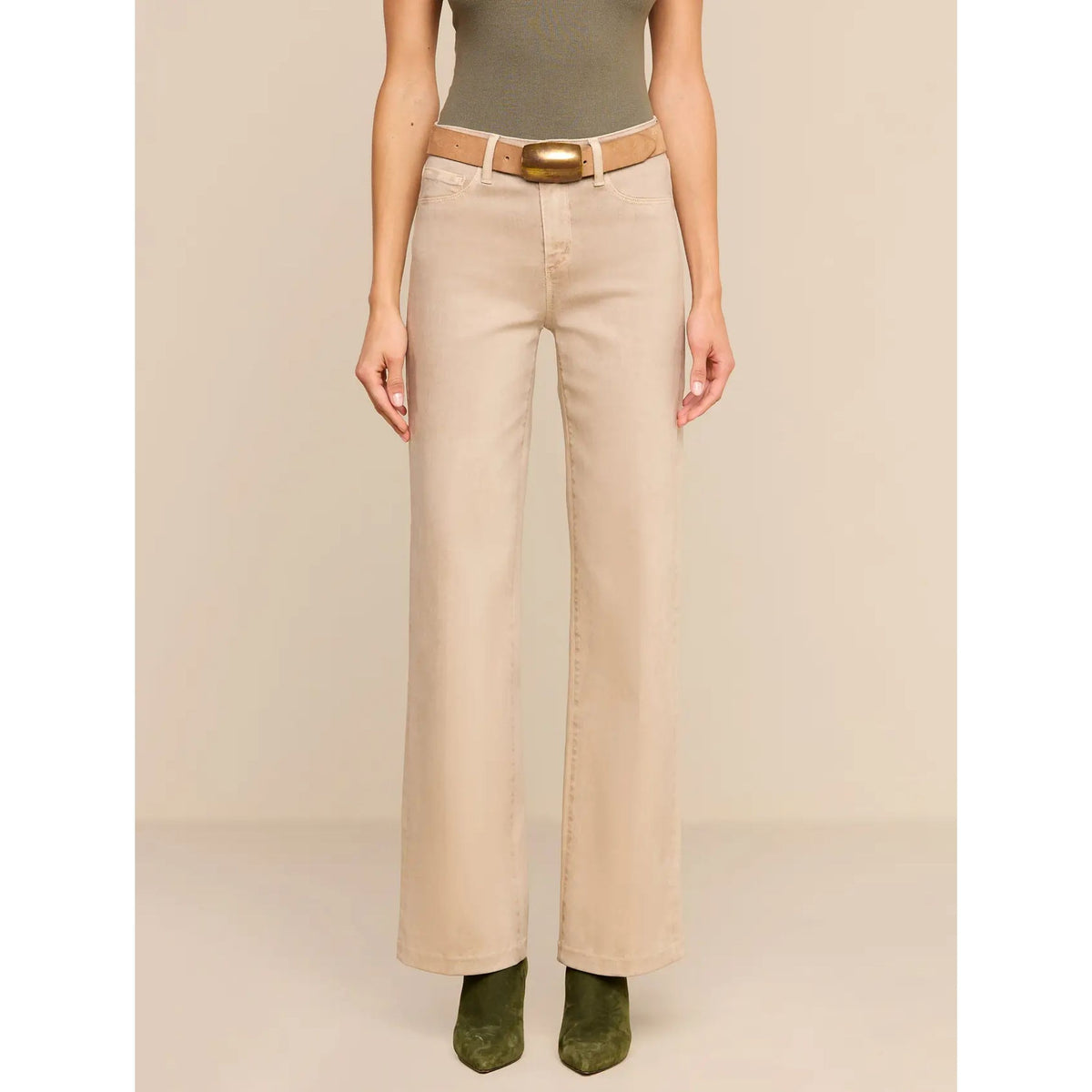 A model is shown from the waist down wearing a pair of straight-leg, wide-leg beige jeans with a tan belt and gold buckle, paired with olive green suede boots against a solid beige studio background.