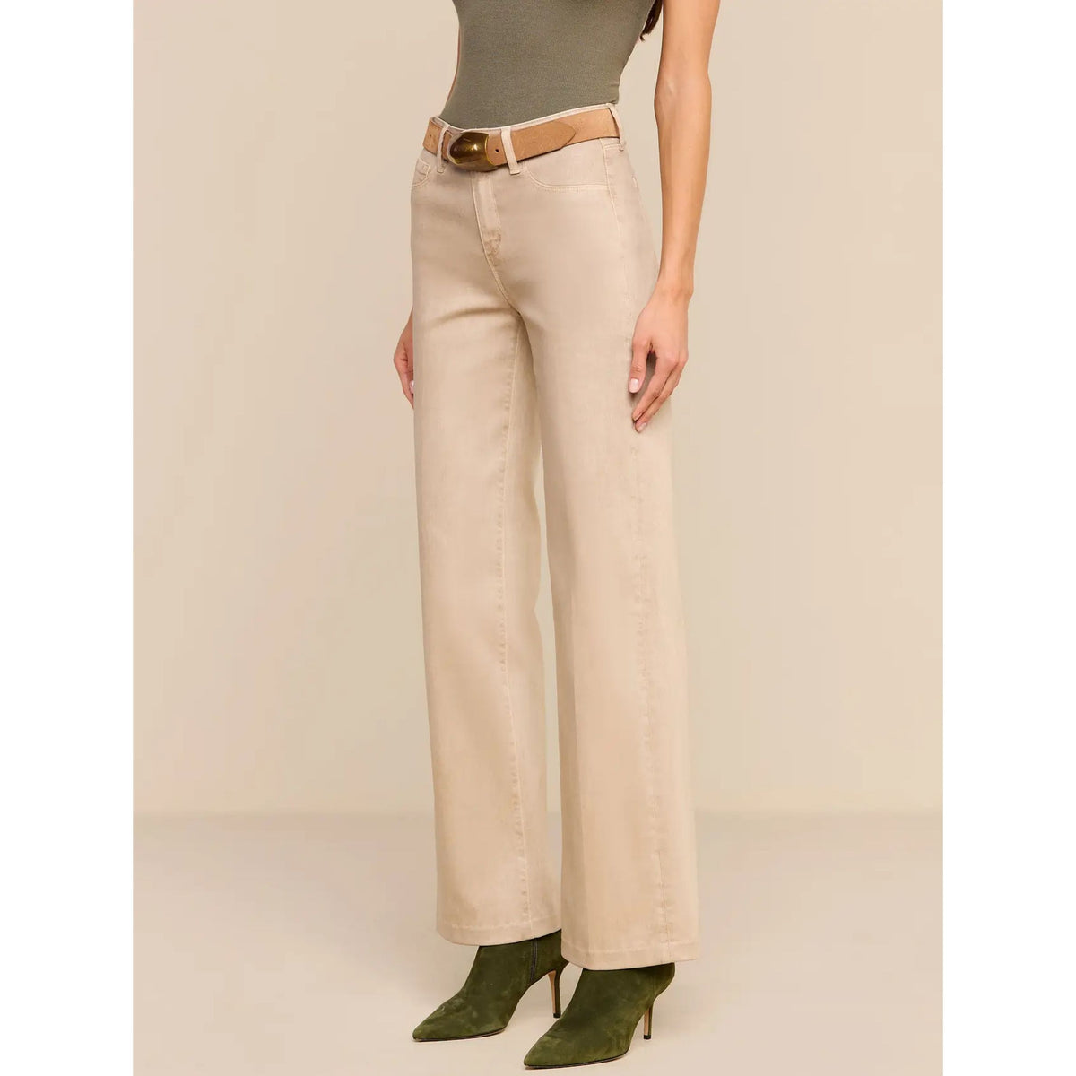 A model wearing beige wide-leg jeans with a tan belt, paired with olive green pointed-toe heels, shown against a solid beige studio background.