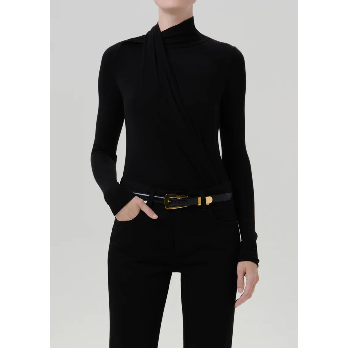 A model wearing a black long-sleeve mock neck top with a draped crossover detail at the collar, paired with black pants and a black belt with a gold buckle, against a light gray studio background.