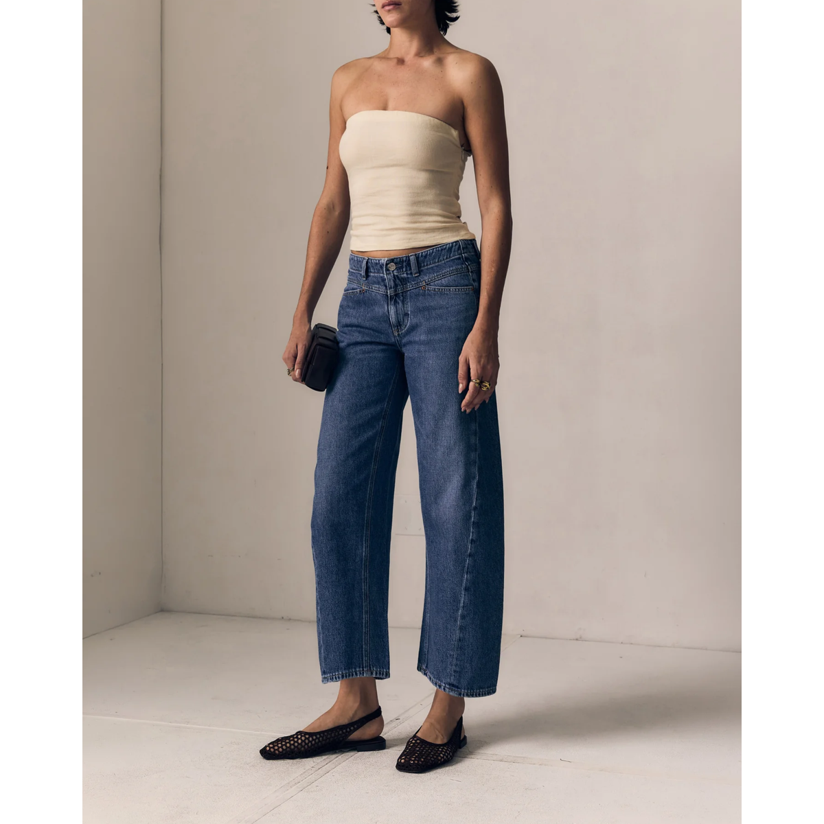 A model wearing a beige strapless tube top, wide-leg cropped blue jeans, and black flat shoes, holding a small black clutch in a minimalist beige room.
