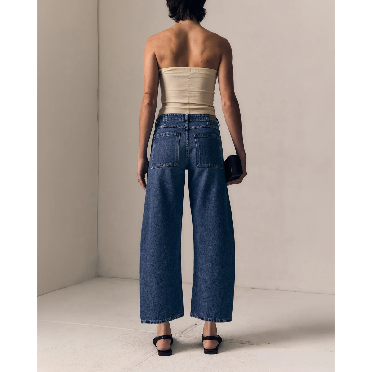 A model viewed from behind wearing high-waisted, wide-leg blue denim jeans, a beige strapless tube top, and black sandals in a minimalist beige room.