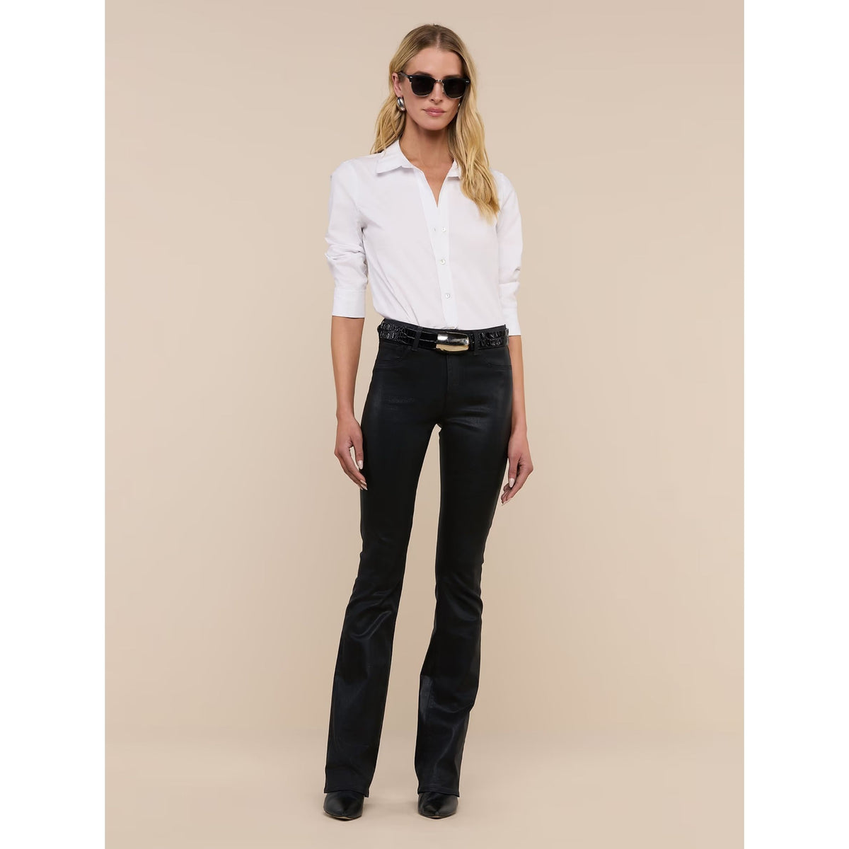 A woman modeling a white long-sleeve button-up shirt and black flared jeans with a black belt, standing against a solid beige studio background.