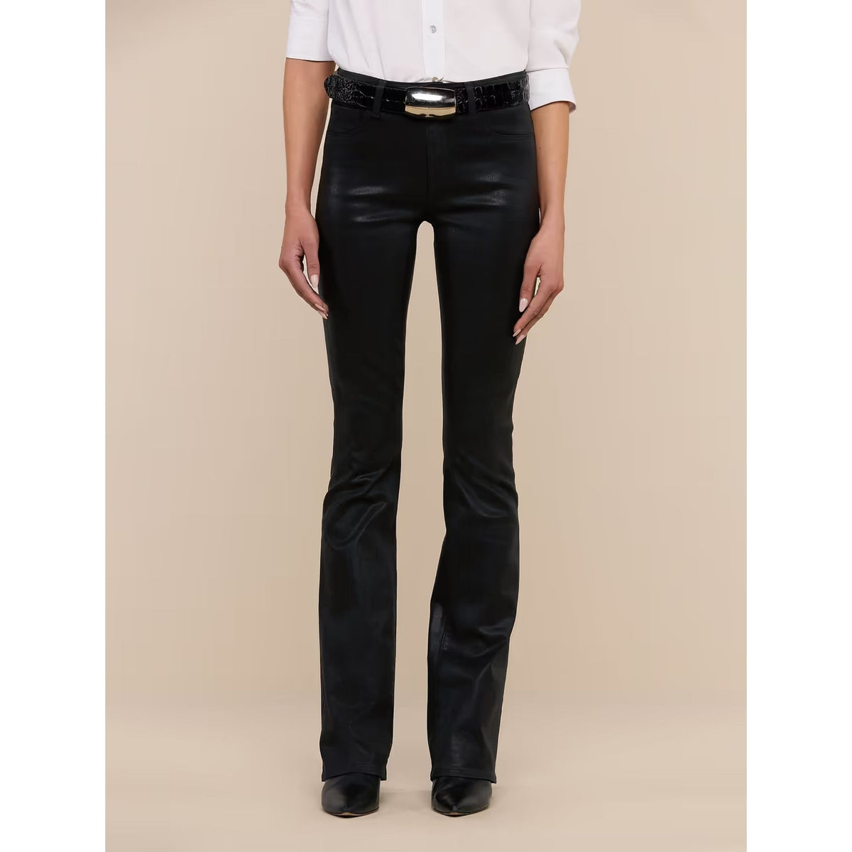 A model wearing black leather flare pants with a black belt featuring a gold buckle, paired with a white button-down shirt and black shoes, set against a solid beige background.