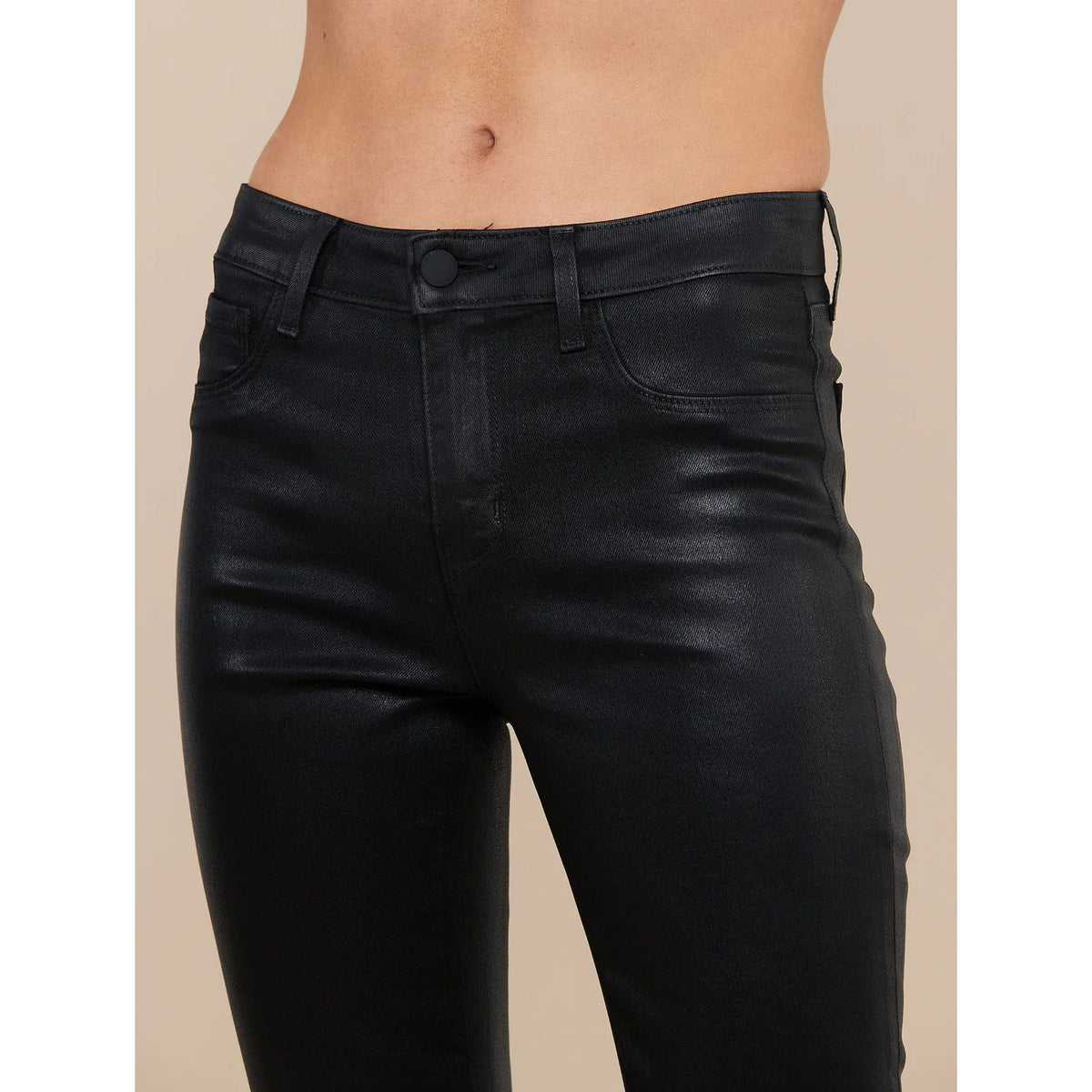 Close-up of a model wearing black faux-leather skinny jeans, showing the front waistband, button, belt loops, and front pockets against a solid beige background.