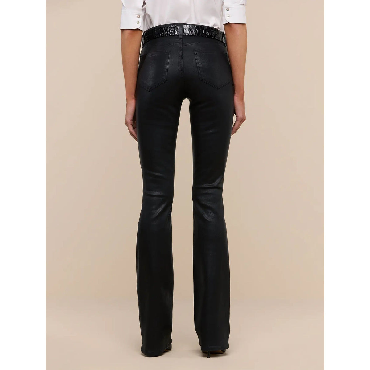 Black leather flare jeans shown from the back on a model, paired with a white shirt and a black sequined belt, against a solid beige background.