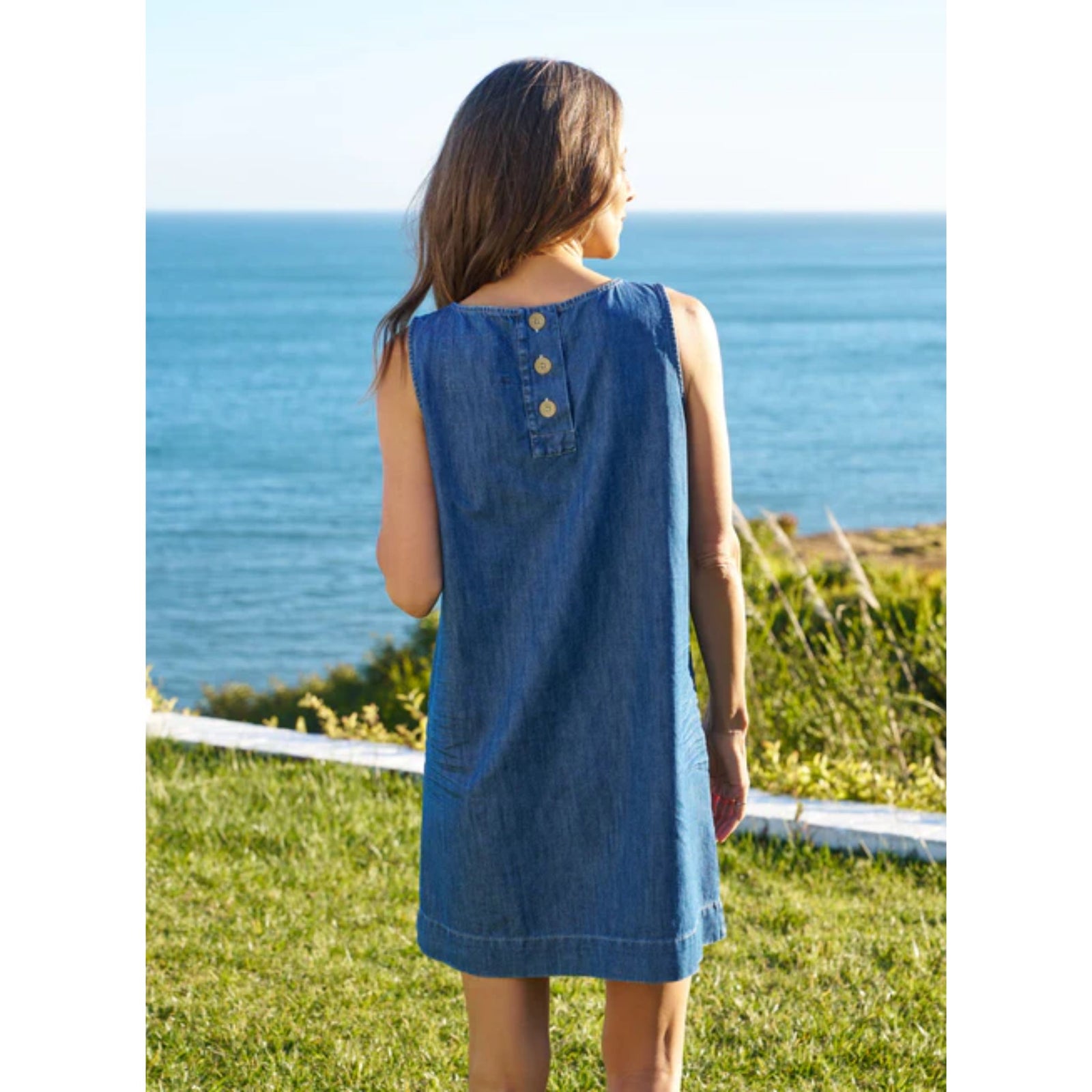 Full-length front view of a model wearing the Frank & Eileen Sheila Shell Dress in Distressed Indigo Wash—a sleeveless denim shift dress with a jewel neckline—standing outdoors against a coastal ocean backdrop.
