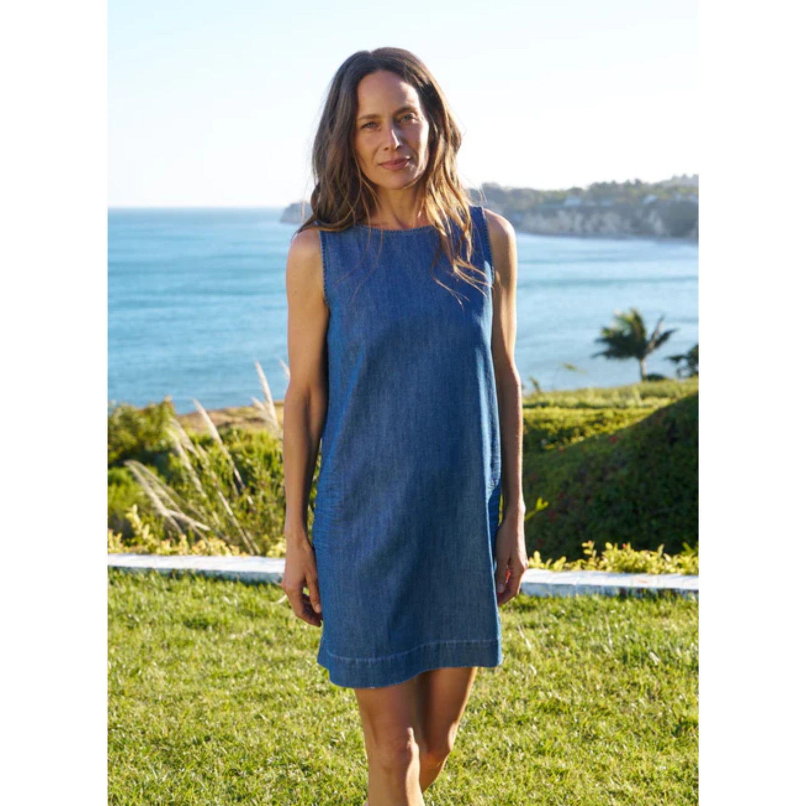 Full-length front view of a model wearing the Frank & Eileen Sheila Shell Dress in Distressed Indigo Wash—a sleeveless denim shift dress with a jewel neckline—standing outdoors against a coastal ocean backdrop.