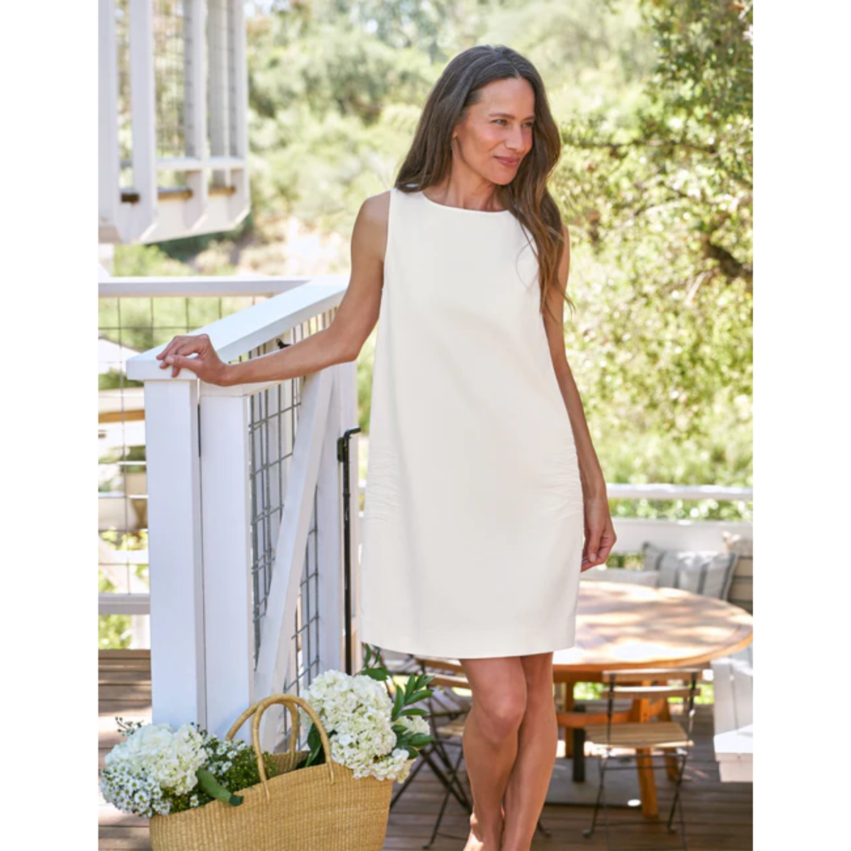A woman wearing a sleeveless cream-colored shift dress stands on a wooden deck, leaning on a white railing with a basket of white flowers in the foreground and a garden background.