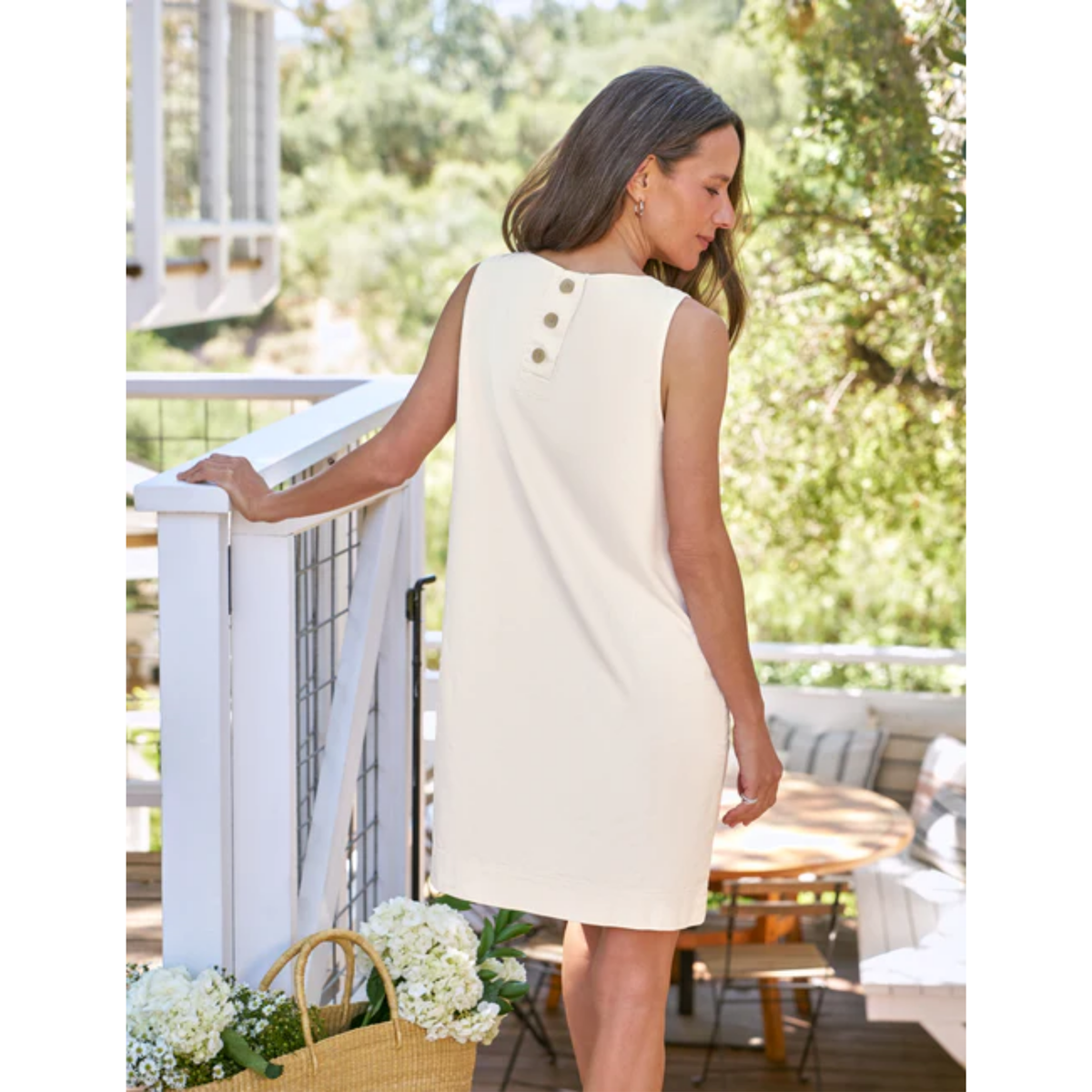 A woman wearing a sleeveless cream-colored dress with three gold buttons on the back, standing on a white wooden deck with a garden background.
