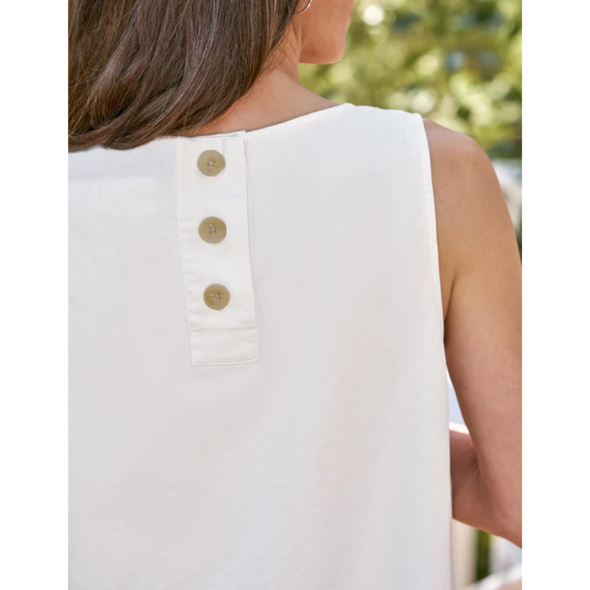 Close-up back view of a woman wearing a sleeveless white top with three gold buttons on the back, set against a blurred outdoor background.