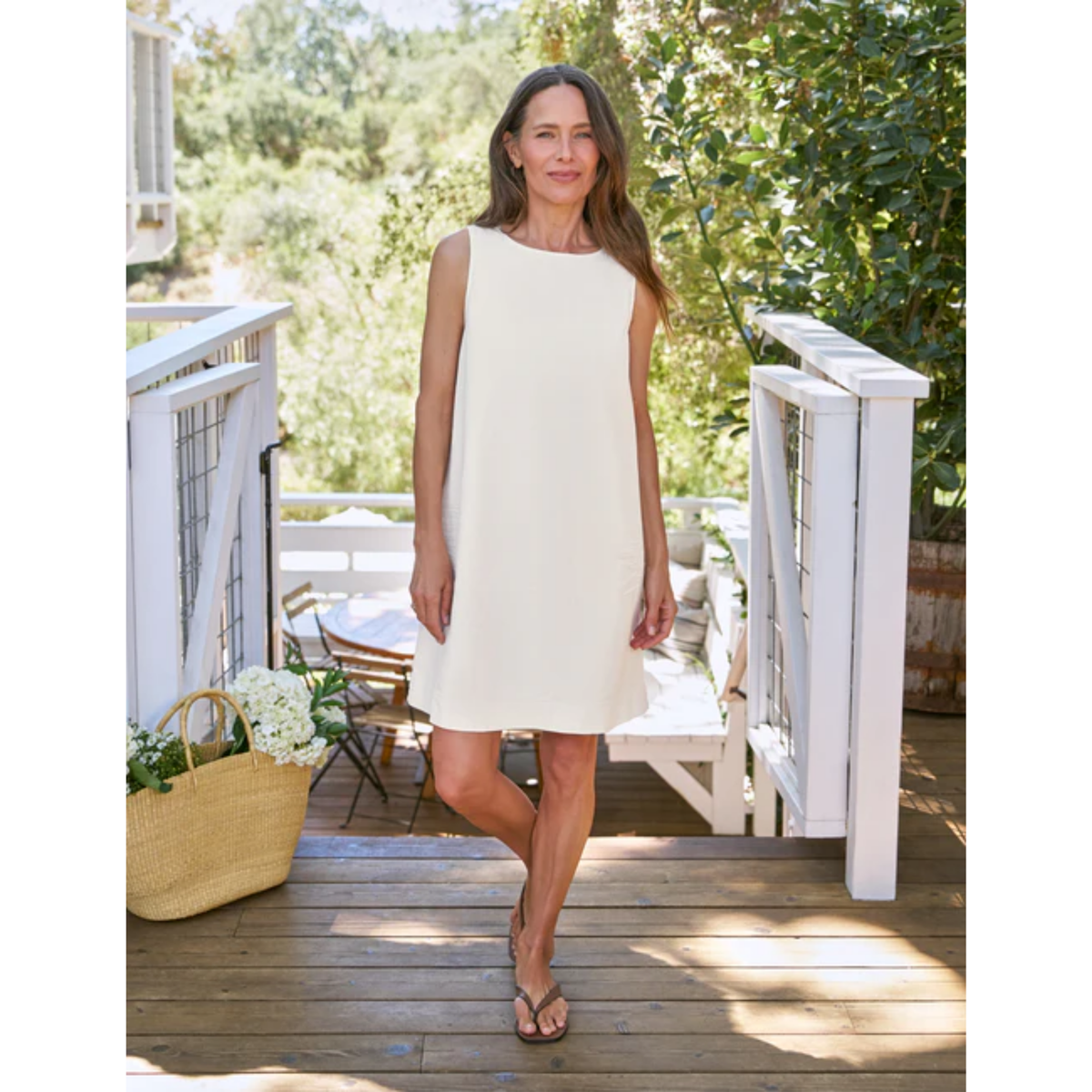 A woman stands on a wooden deck wearing a sleeveless cream-colored shift dress and flat sandals, with white railings and green foliage in the background.
