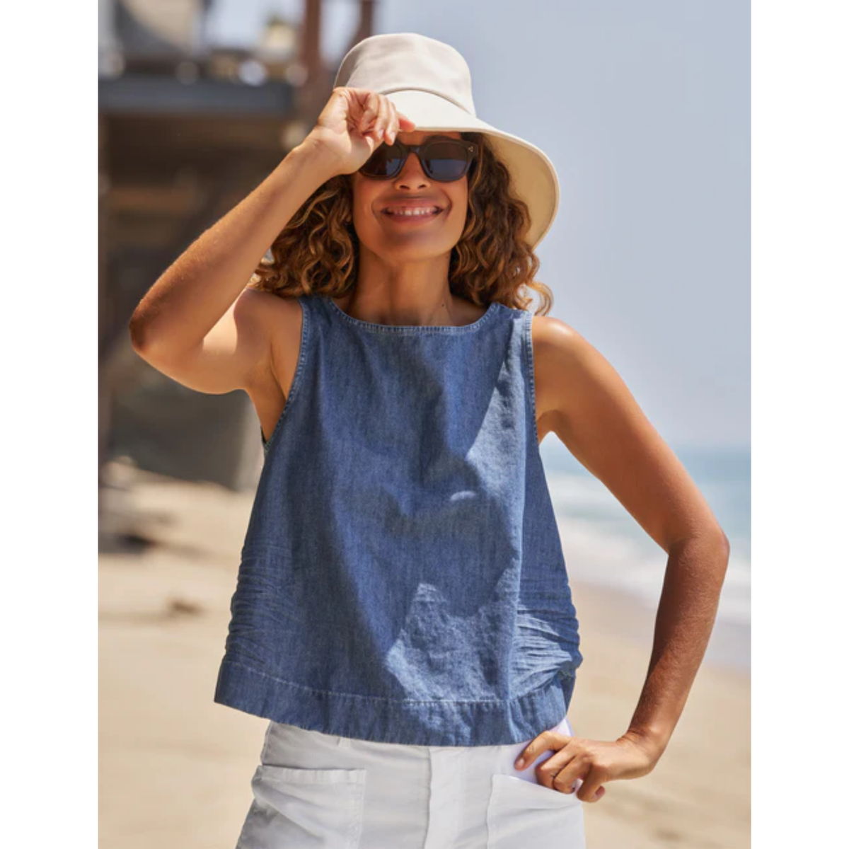 A woman wearing a sleeveless blue denim-style top, white pants, a beige bucket hat, and sunglasses stands on a beach with the ocean in the background.