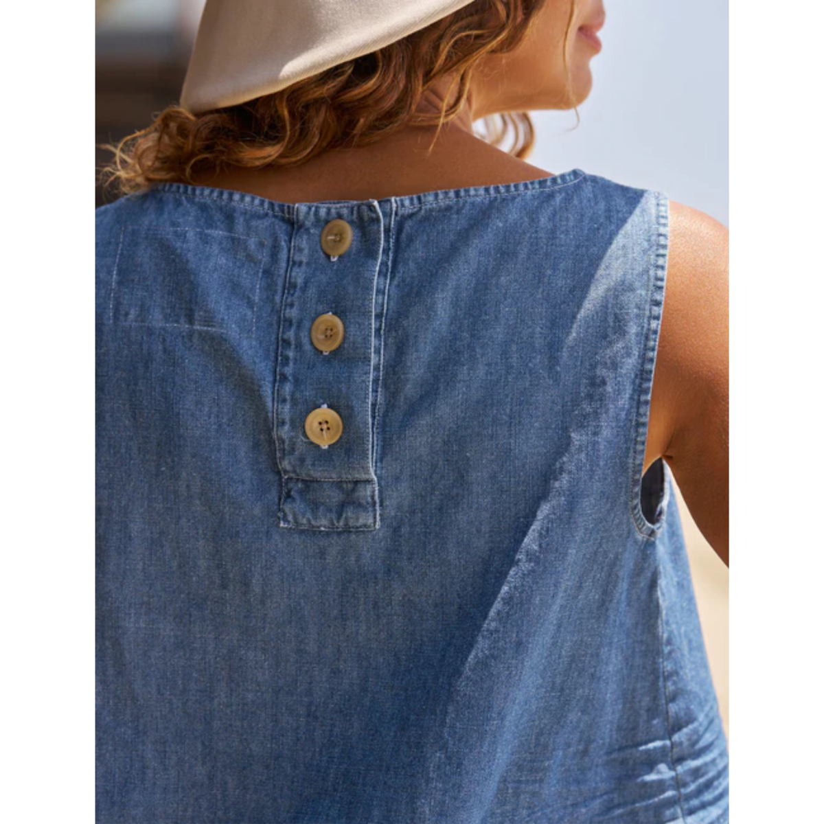 Close-up back view of a woman wearing a sleeveless blue denim top with three decorative buttons at the neckline, shown outdoors against a bright sky.