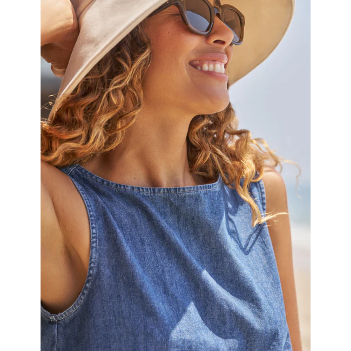 A woman wearing a sleeveless blue denim top, a beige wide-brim hat, and sunglasses, photographed outdoors against a bright sky.