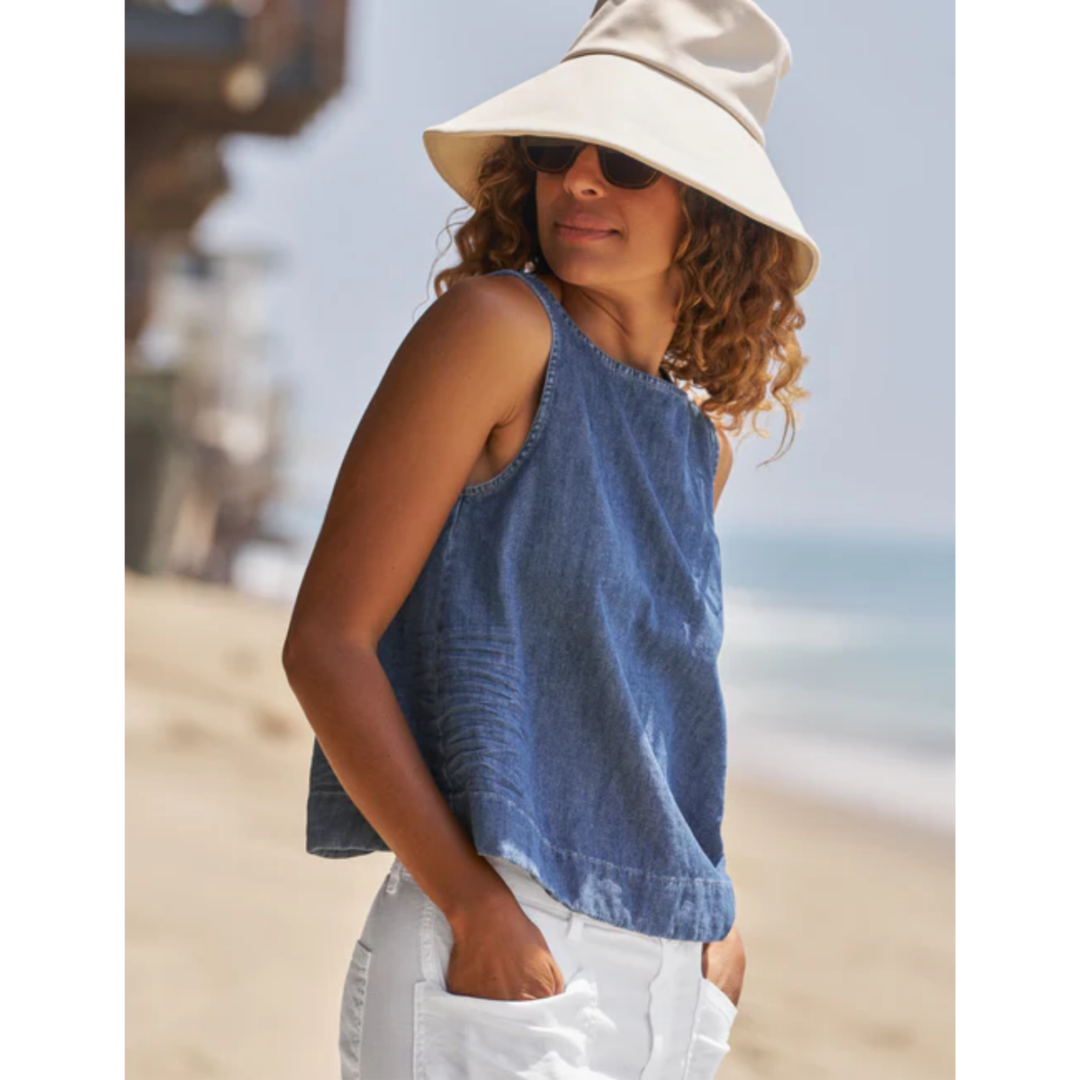 A woman on a beach wearing a sleeveless blue denim top, white pants, and a large cream sun hat.