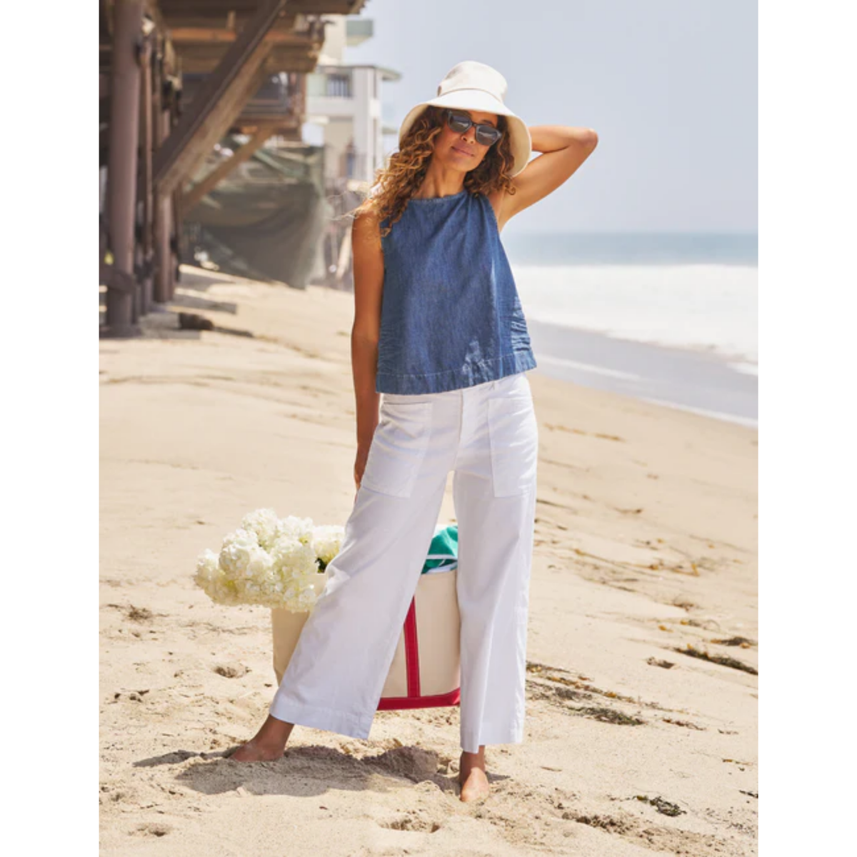 A woman stands on a sandy beach wearing a sleeveless blue top, white wide-leg pants, a white hat, and sunglasses, with a beach umbrella and flowers beside her.