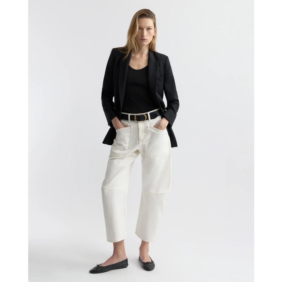 A model stands against a light gray studio background wearing a black blazer, black top, white cropped pants with a black belt, and black flats.