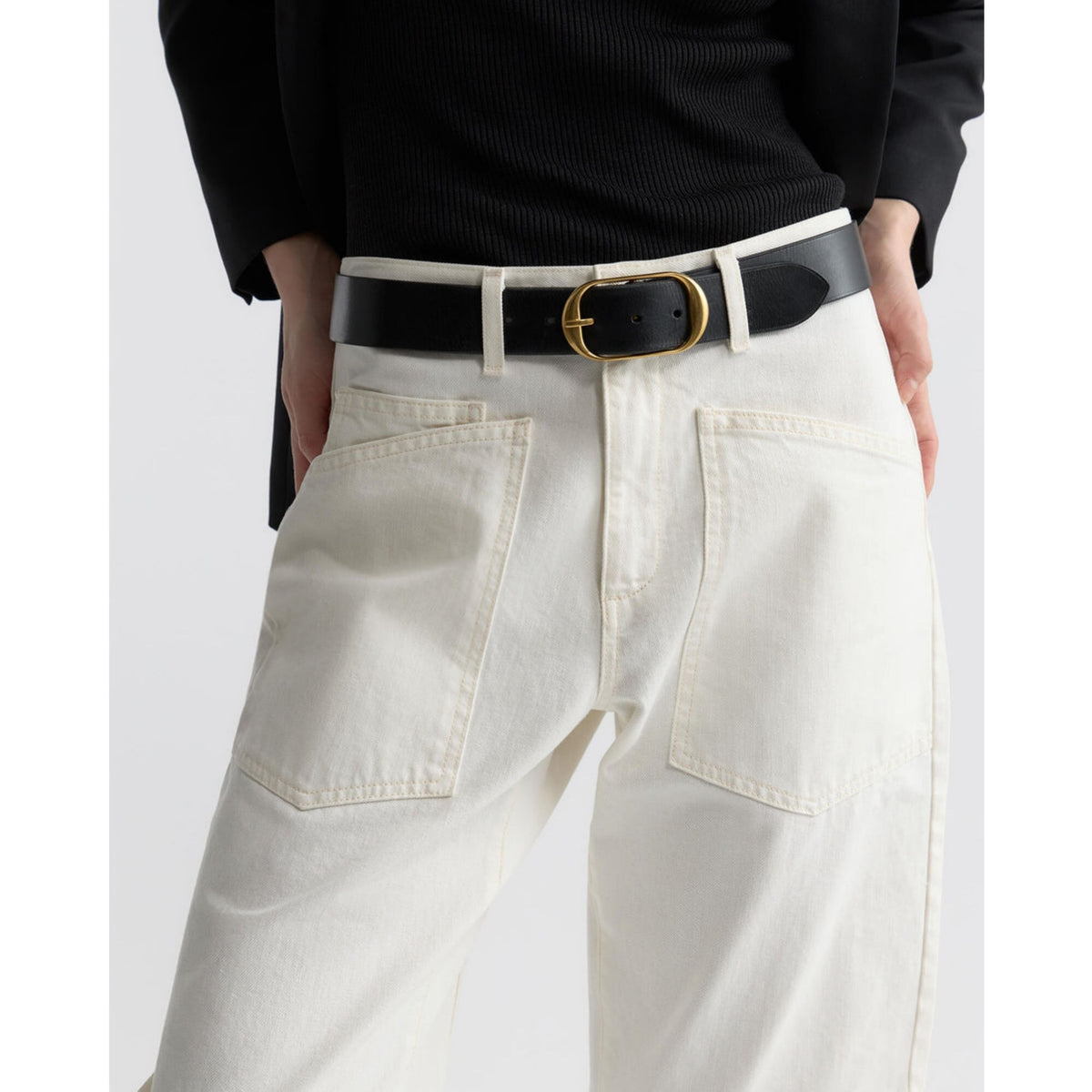 A close-up of a person wearing off-white wide-leg jeans with a black leather belt featuring a gold-tone oval buckle, paired with a black ribbed top.