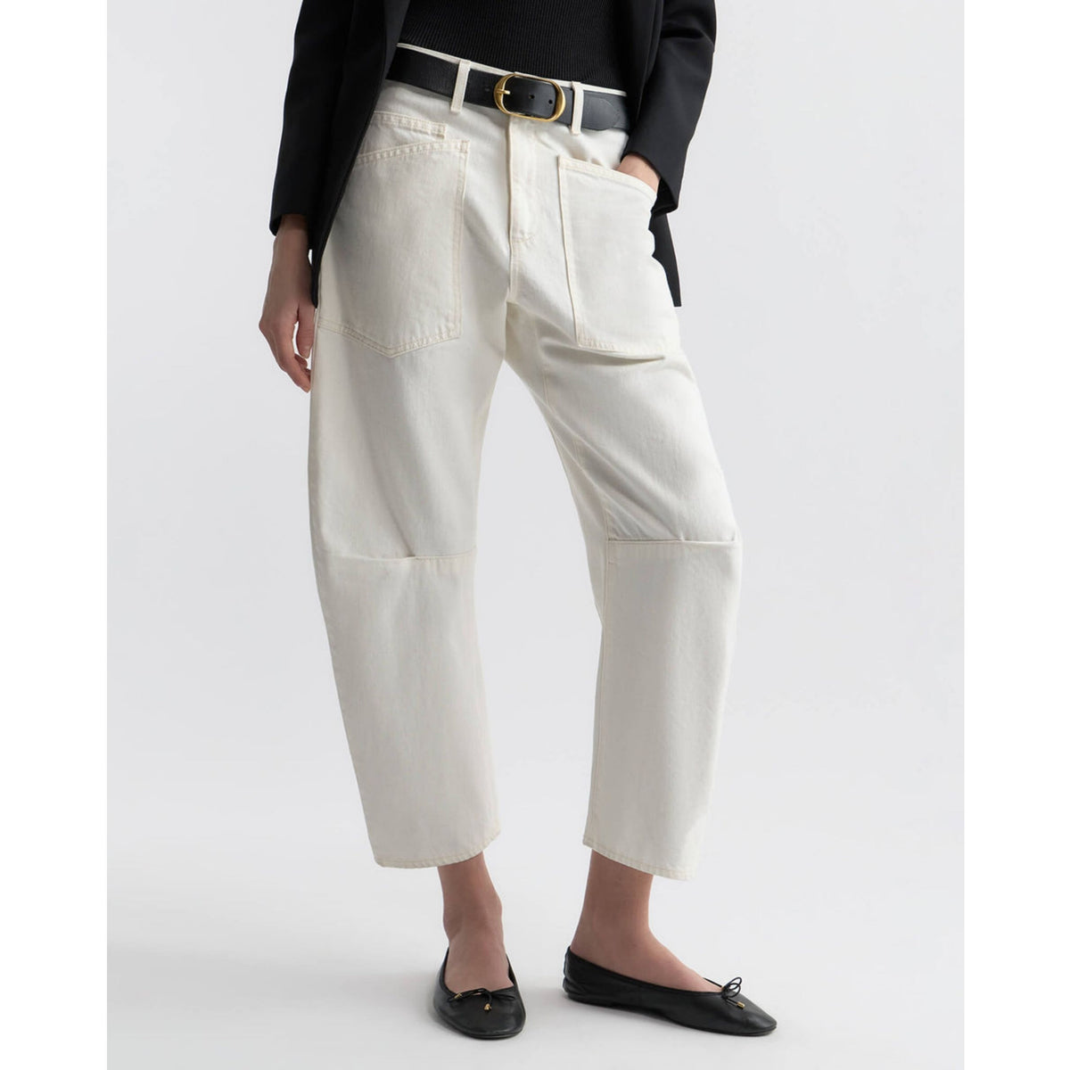 A model is shown from the waist down wearing off-white cropped trousers with patch pockets and knee patches, styled with a black belt, a black blazer, and black flat shoes against a light gray background.
