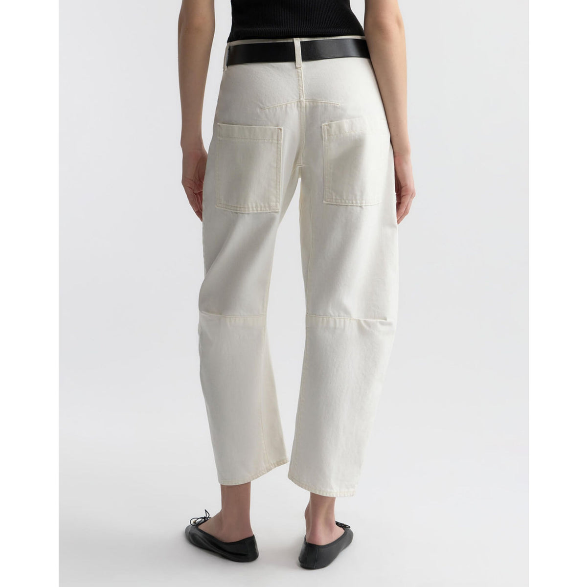 A model shown from the back wearing off-white cropped denim pants with a black belt, black top, and black flat shoes against a light gray studio background.