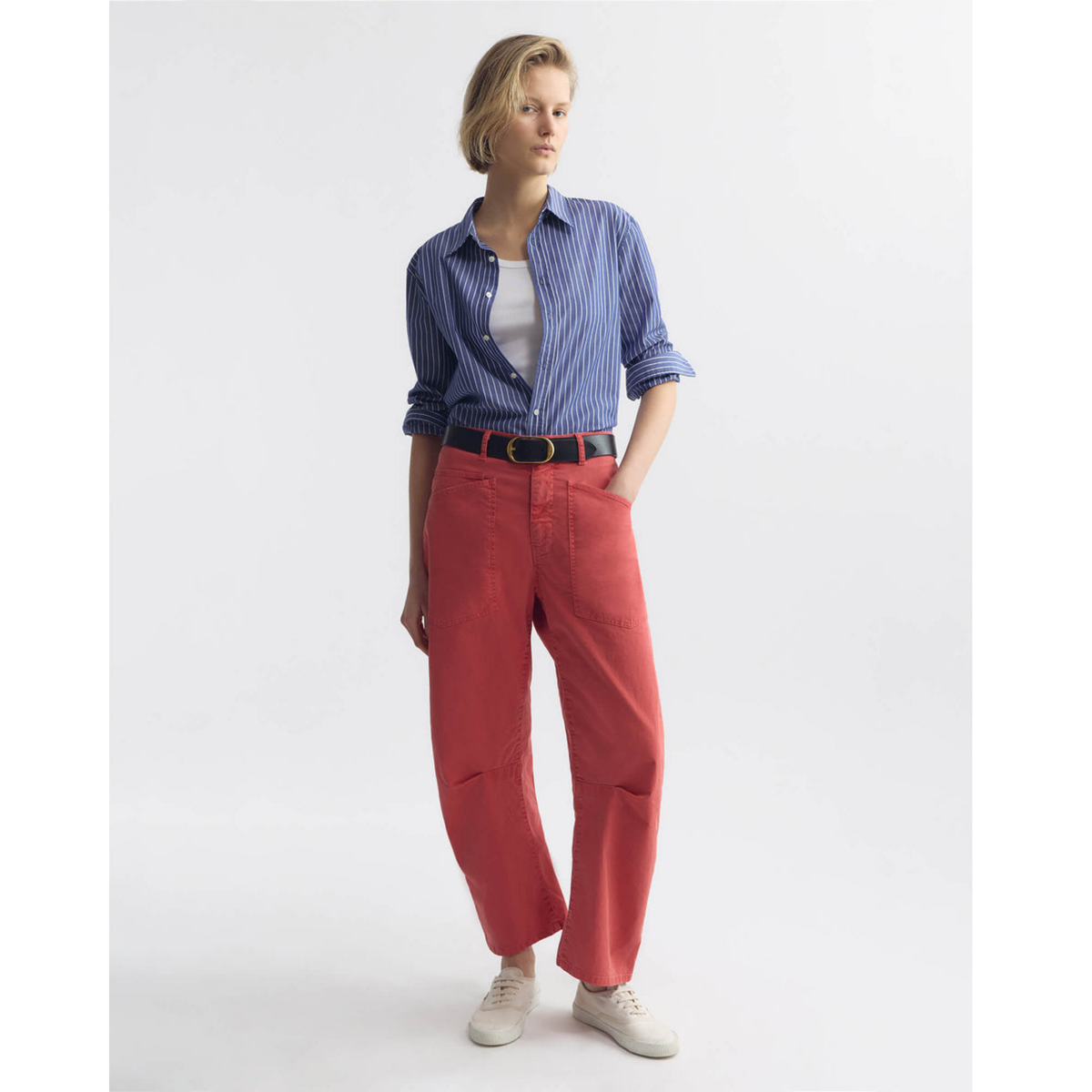 Woman wearing a blue striped shirt, white top, red pants, and white sneakers on a light gray background