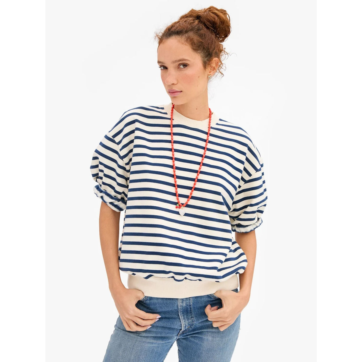 Front view of a model wearing the short-sleeve "Oversized Sweatshirt" in cream and navy horizontal stripes, featuring a ribbed crew neck and hem, paired with blue denim jeans and an orange bead necklace.